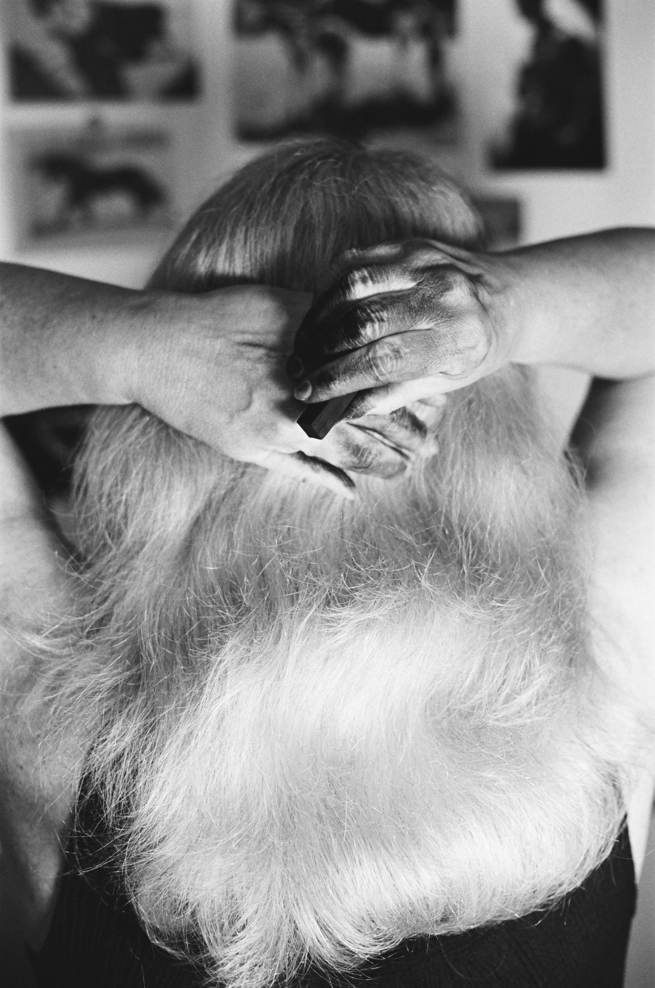 A person with a long white beard and hair is getting a haircut, with hands on their head in a barbershop or salon. There are framed pictures on the wall in the background.