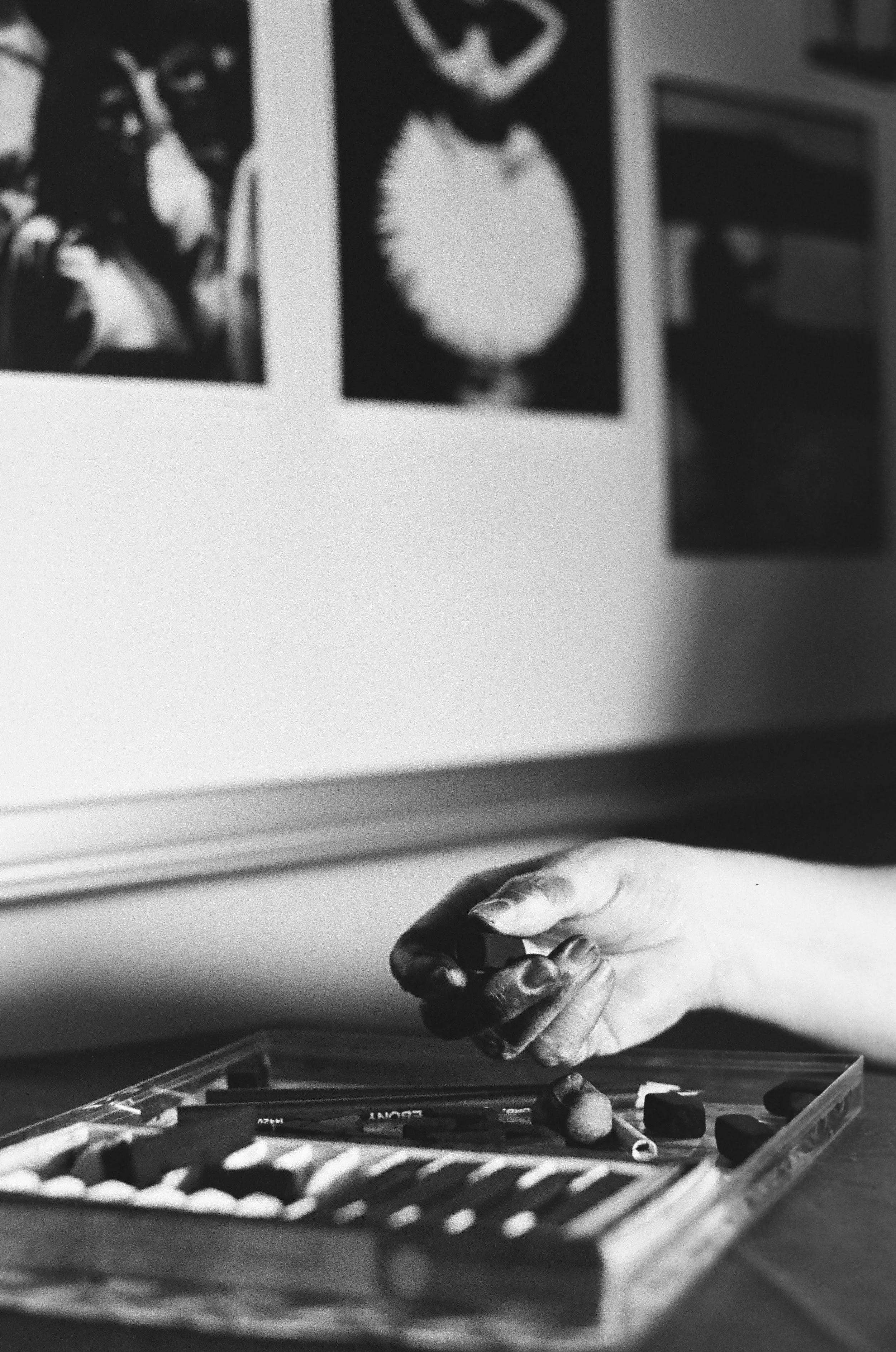A person's hand covered in black chalk resting on a box of chalk and art supplies on a table, with artwork displayed on the wall in the background.