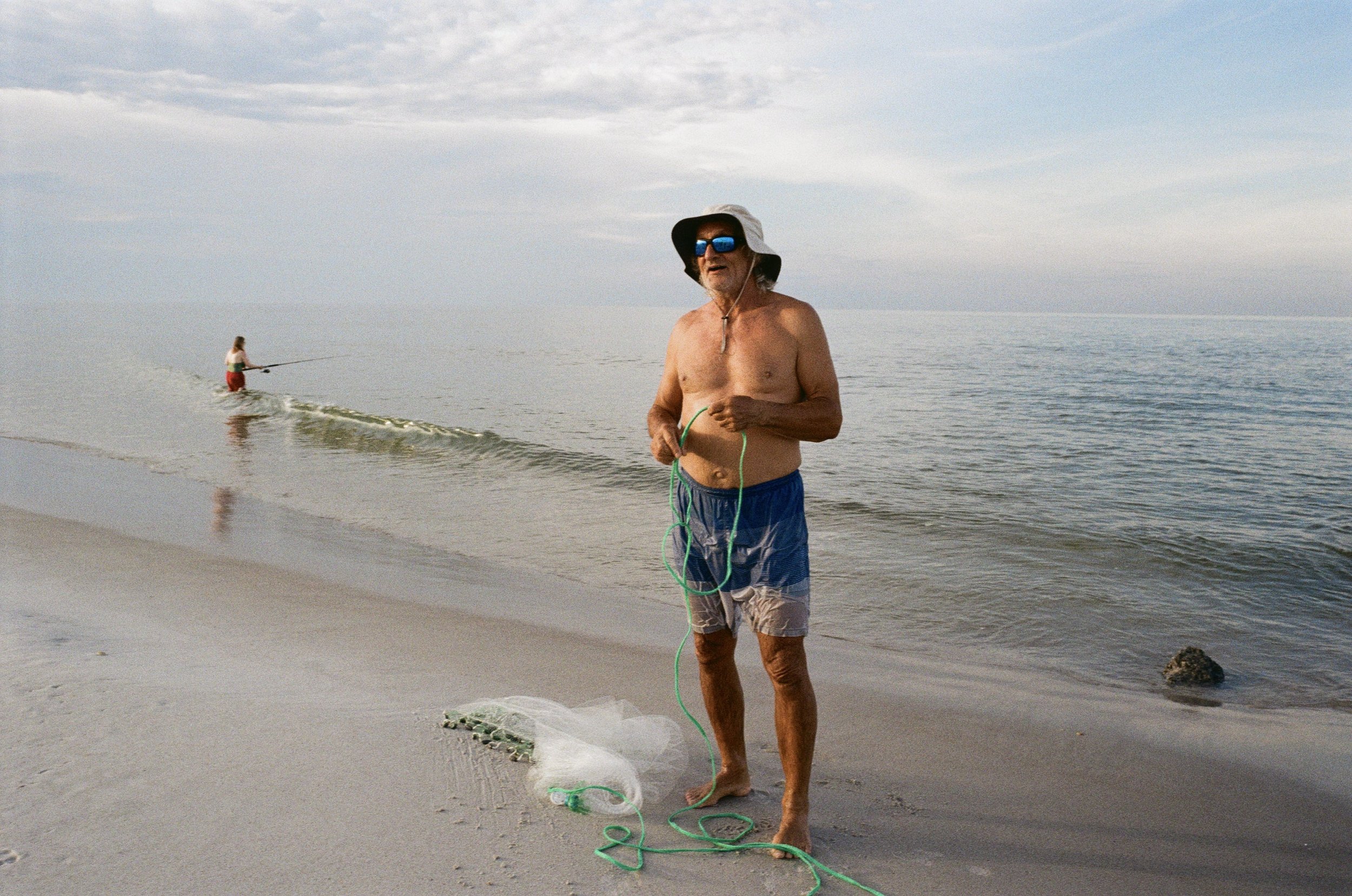 An elderly man in blue swim trunks, wearing sunglasses and a wide-brimmed hat, standing on a sandy beach holding a fishing net with a piece of green rope. In the background, a woman in a white top and red shorts is fishing in the water. The sky is partly cloudy, and the water is calm.
