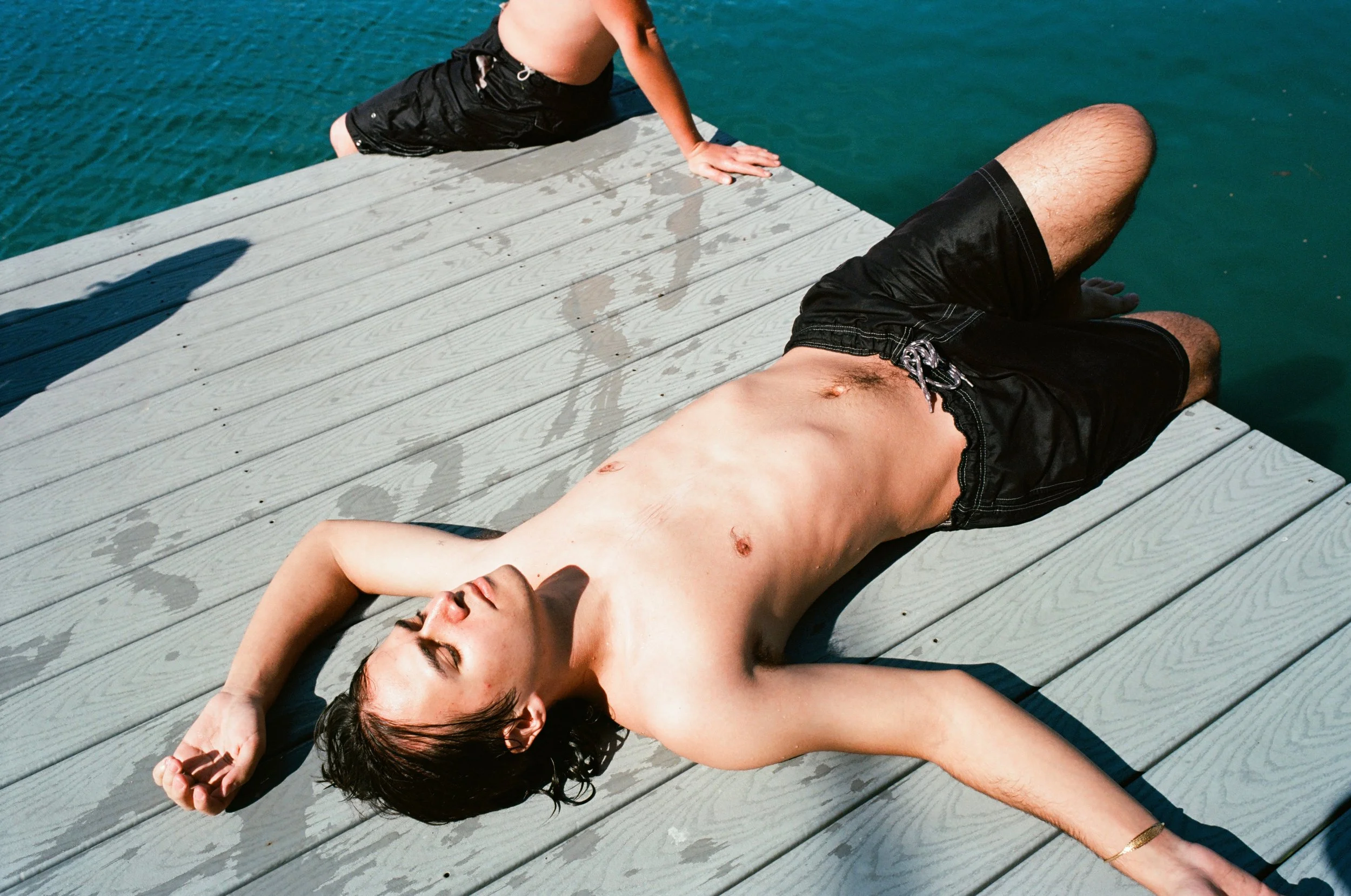 A young man sunbathing on a wooden dock by the water, with only swim shorts on, eyes closed, relaxing in the sunlight.
