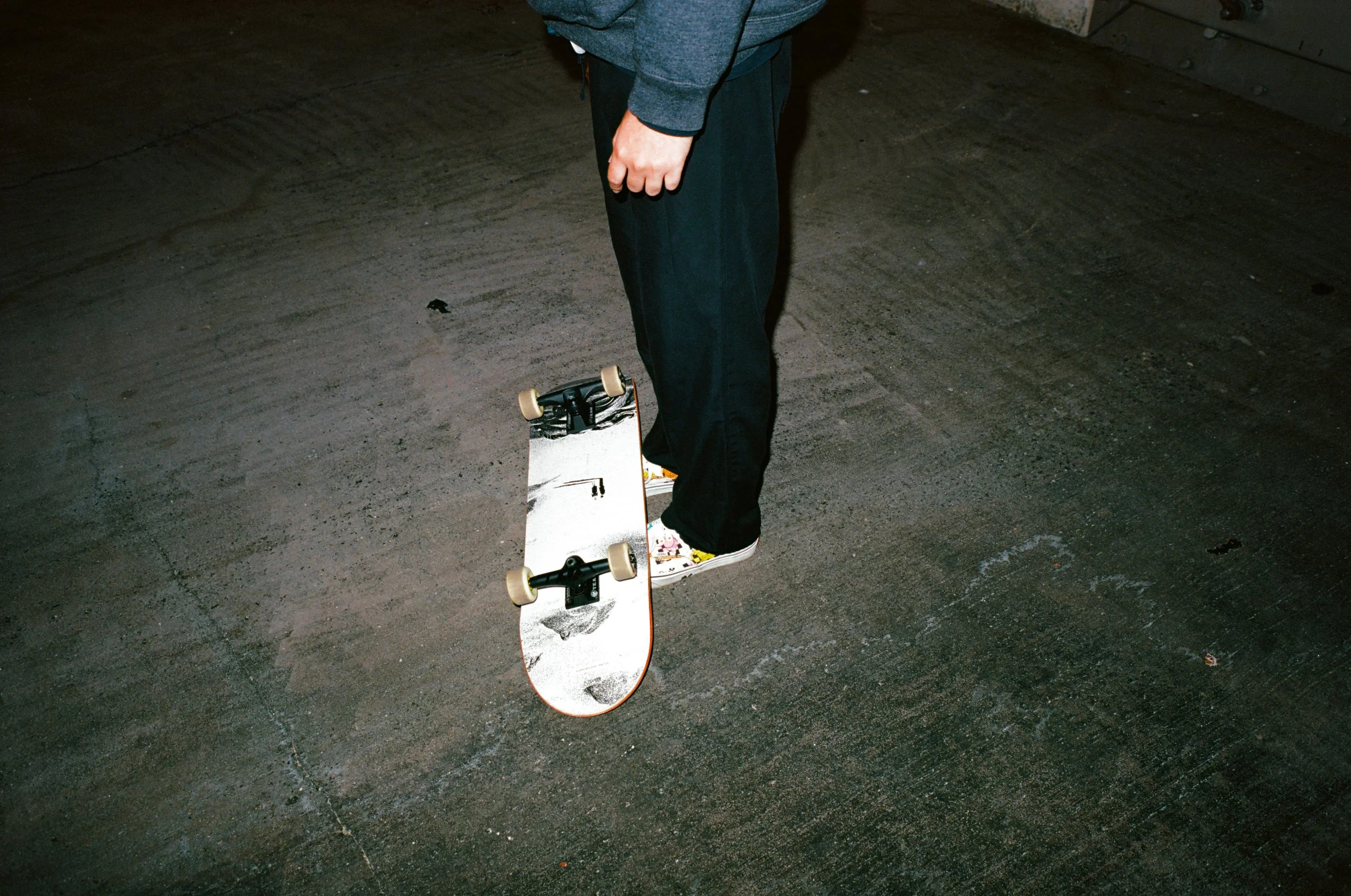 Person standing on skateboard on pavement surface, wearing black pants and a dark hoodie.