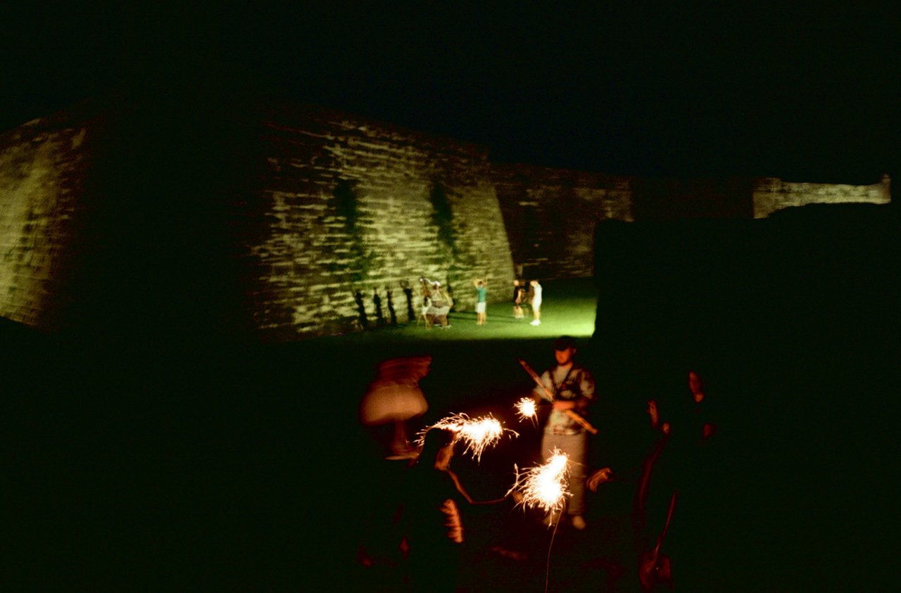 Nighttime scene of a historical fort with people viewing the illuminated stone walls, and children playing with sparklers in the foreground.