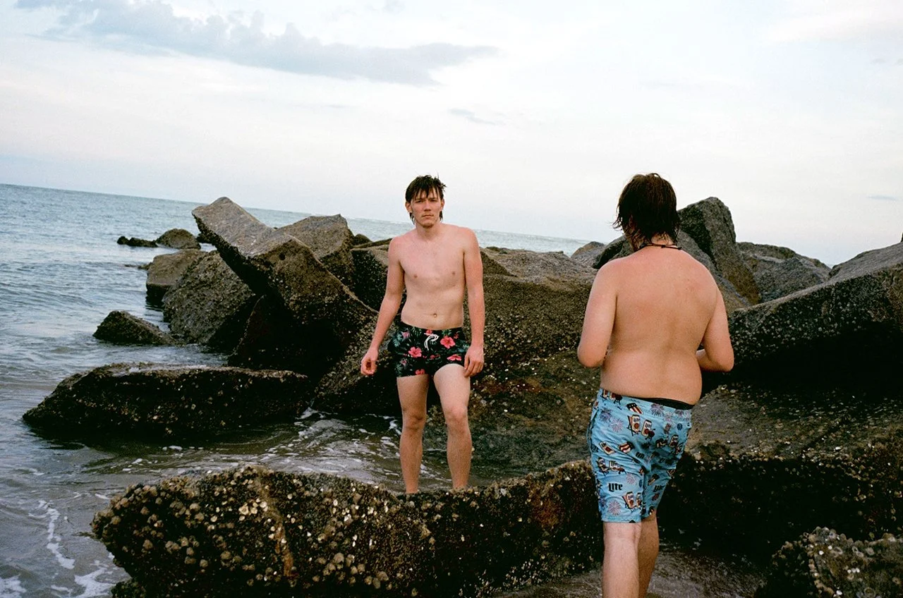 Two young men in swim trunks standing on rocks at the beach, with the ocean and cloudy sky in the background.