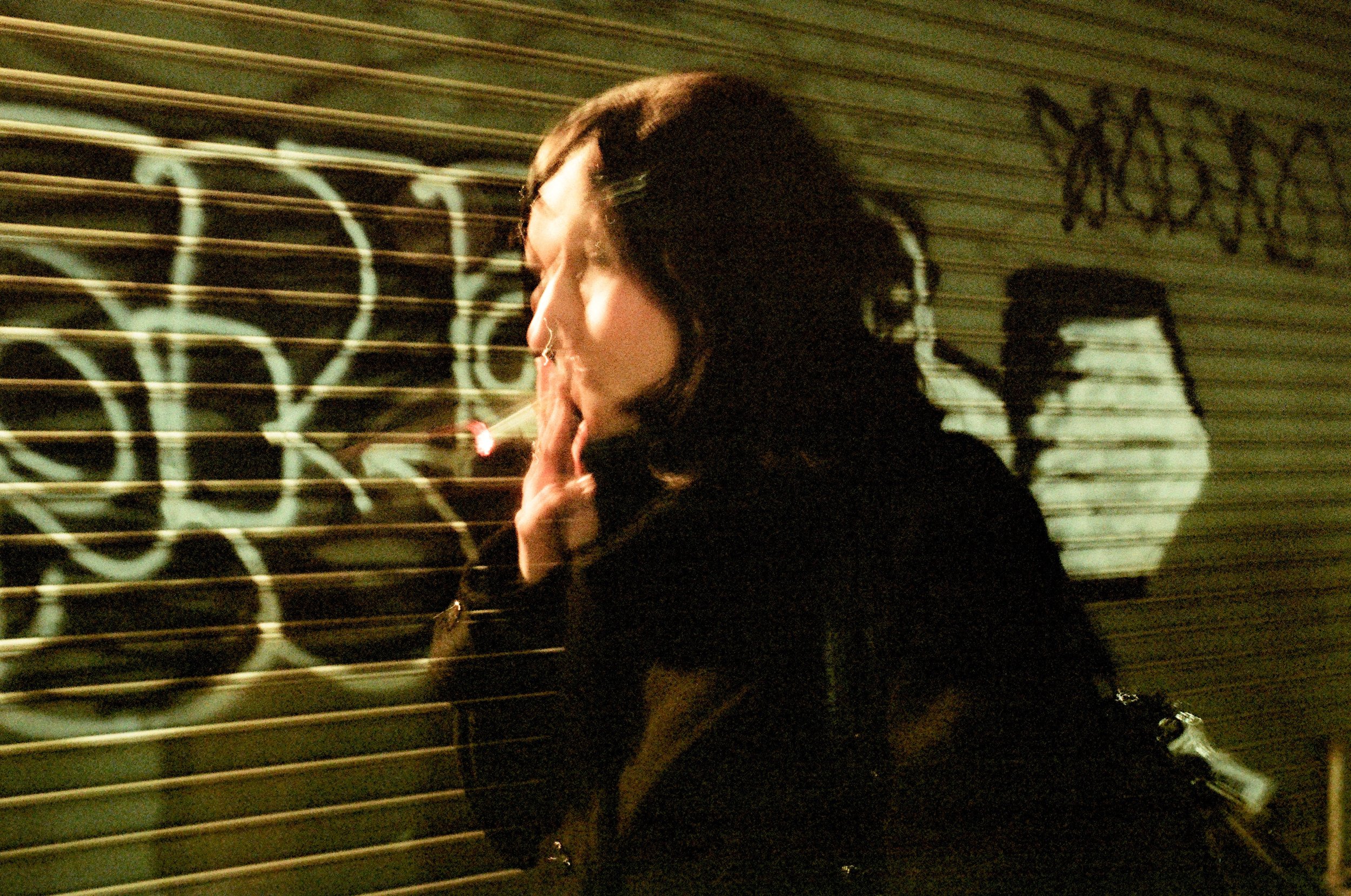 A woman with dark hair, seen in profile, smokes a cigarette in front of a graffiti-covered metal shutter at night.