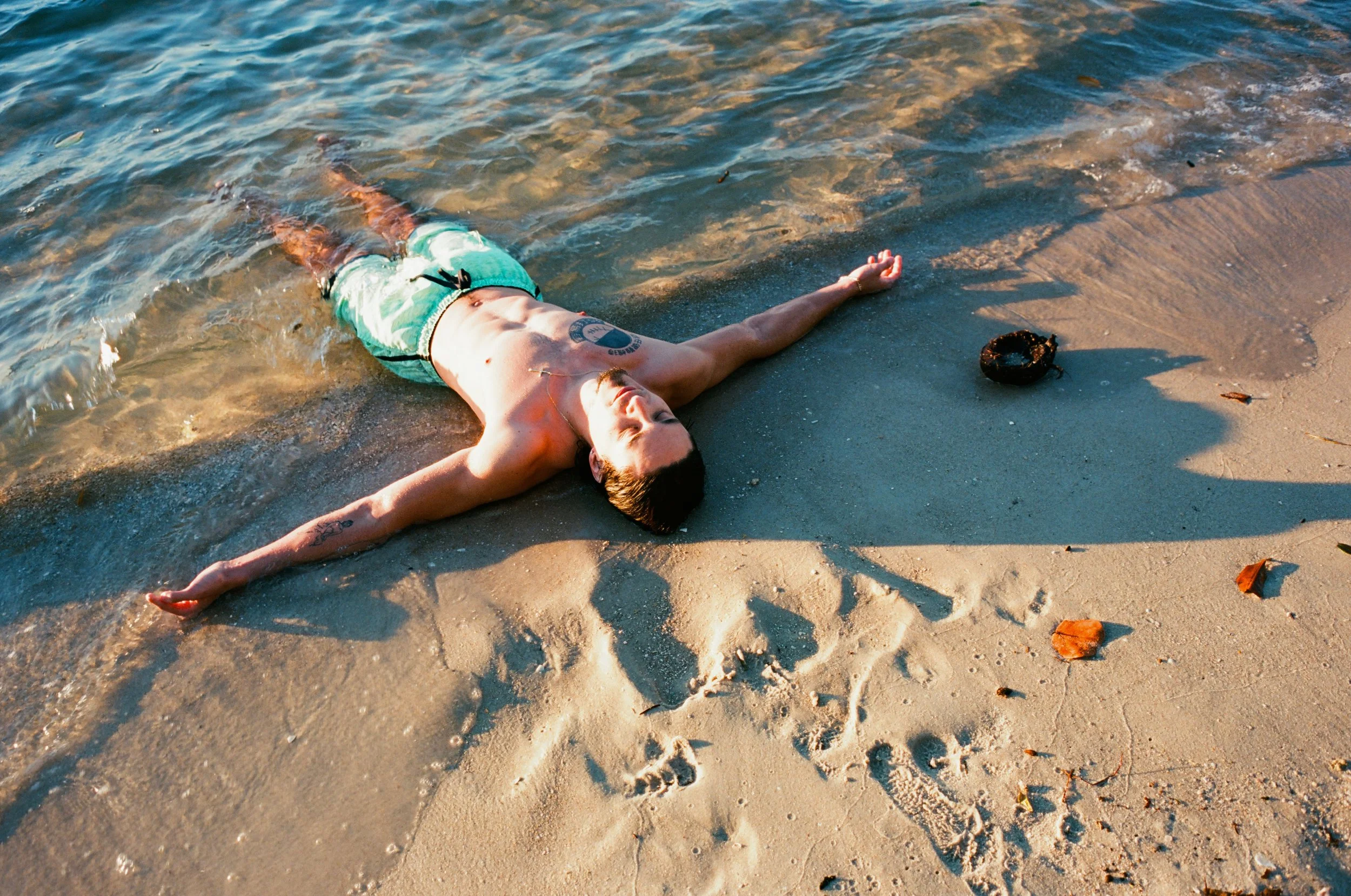 Young man lying on the sandy beach near the water, with arms outstretched and eyes closed, wearing swim shorts and sunglasses. A small, black, round object is nearby on the sand, with footprints and scattered leaves around.