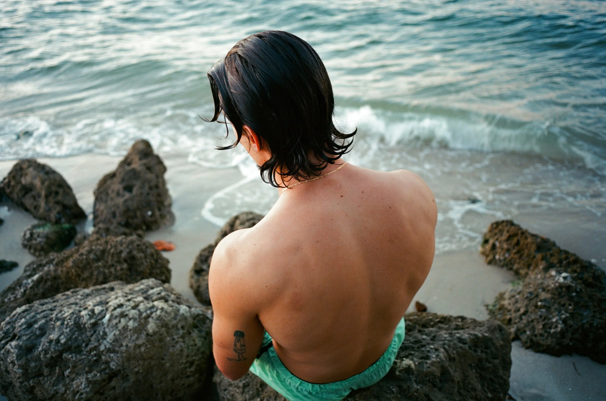 Person with black hair sitting on rocks at the beach, facing the water, with waves coming ashore.