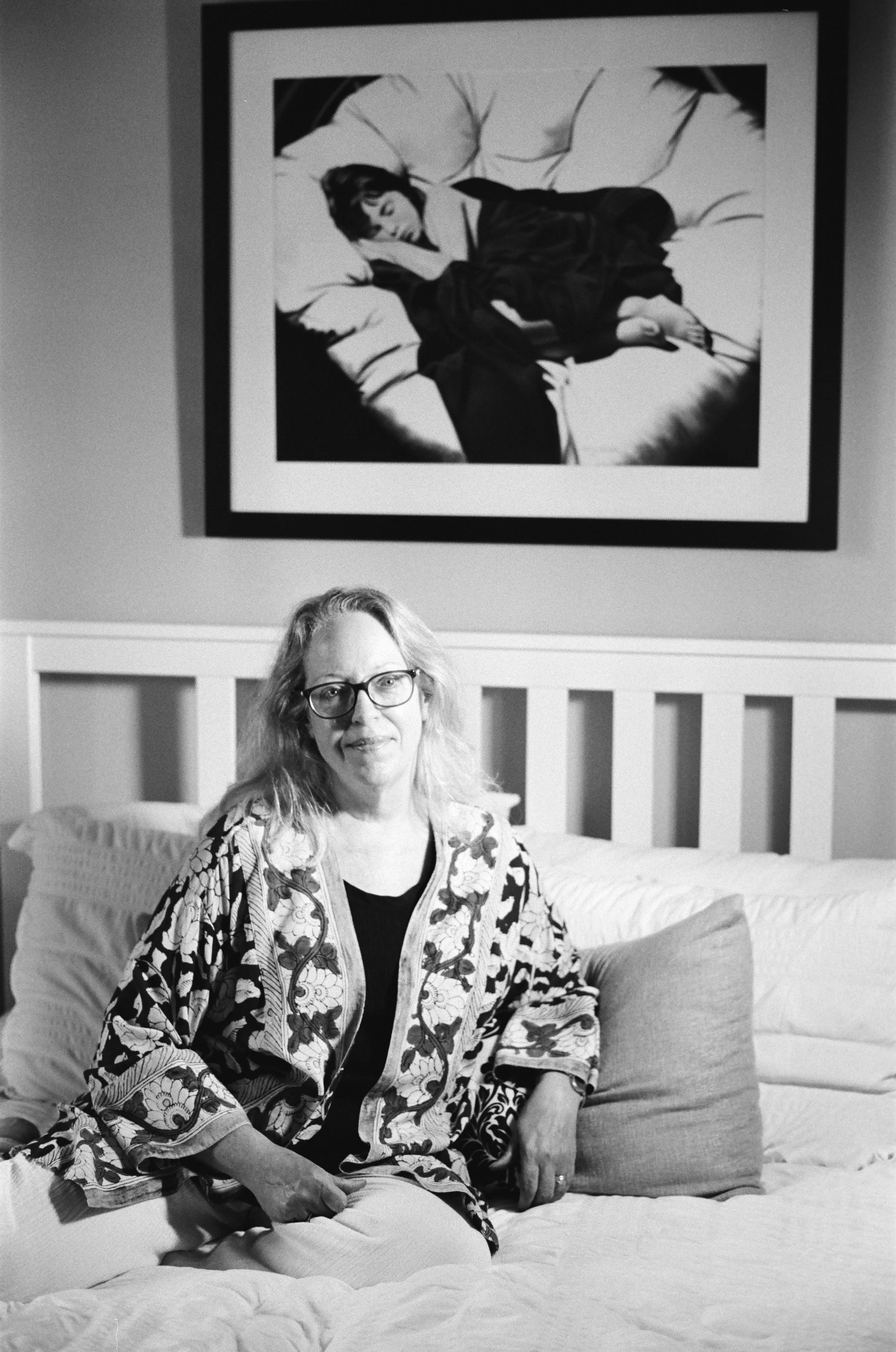 A woman with glasses and wavy hair, wearing a patterned jacket, sitting on a bed in a room, with a painting of a woman sleeping on the wall behind her.