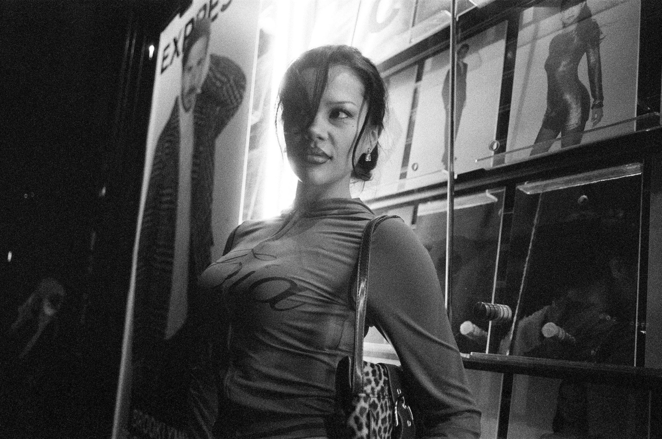 A woman with short dark hair and earrings looking over her shoulder, standing in front of glass display cases with figurines and posters in the background, in a black and white photo.