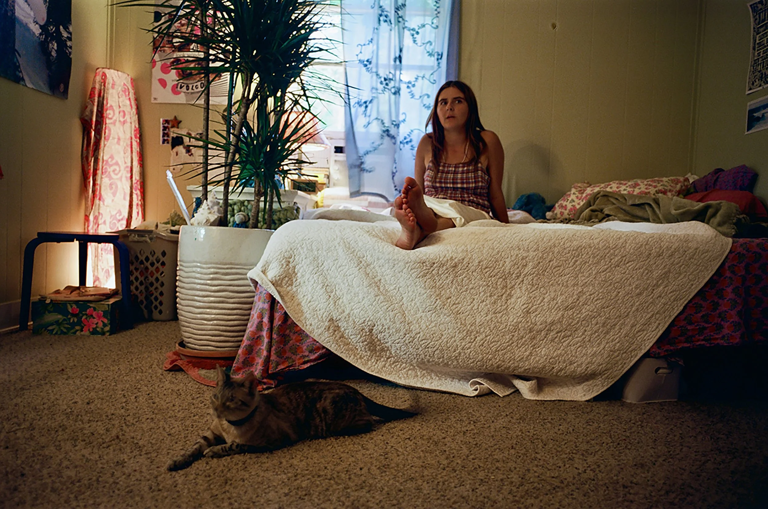 A woman sits on a bed with her legs extended and feet crossed, looking surprised or concerned. A tabby cat is lying on the floor next to the bed. The bedroom has warm lighting, a large potted plant, and various personal items on nearby furniture and shelves, with a window with sheer curtains in the background.