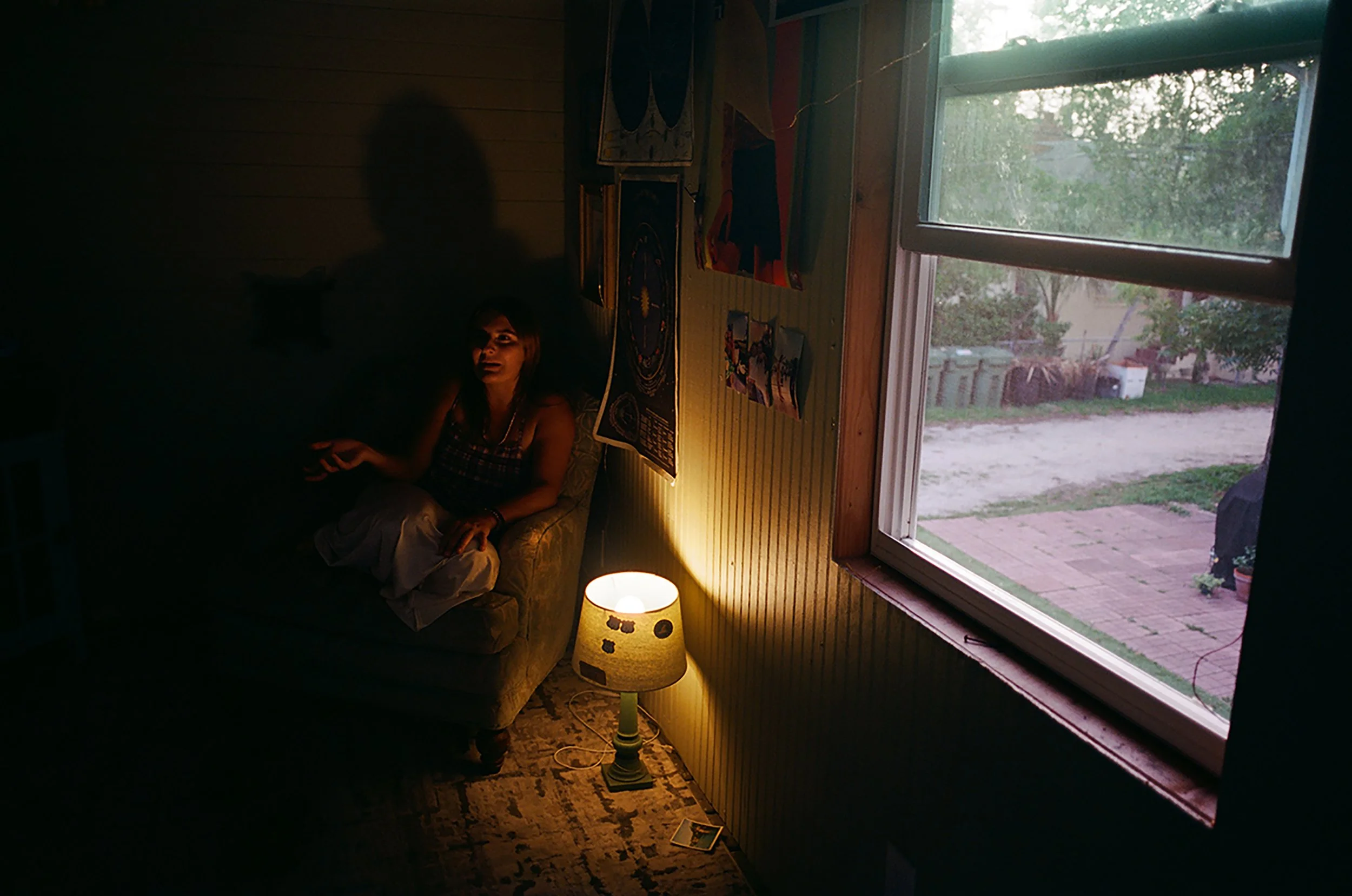A woman sitting on a beige armchair in a dimly lit room with a small lamp on a side table and artwork hanging on the wall. A large window shows an outdoor scene with trees and a patio.
