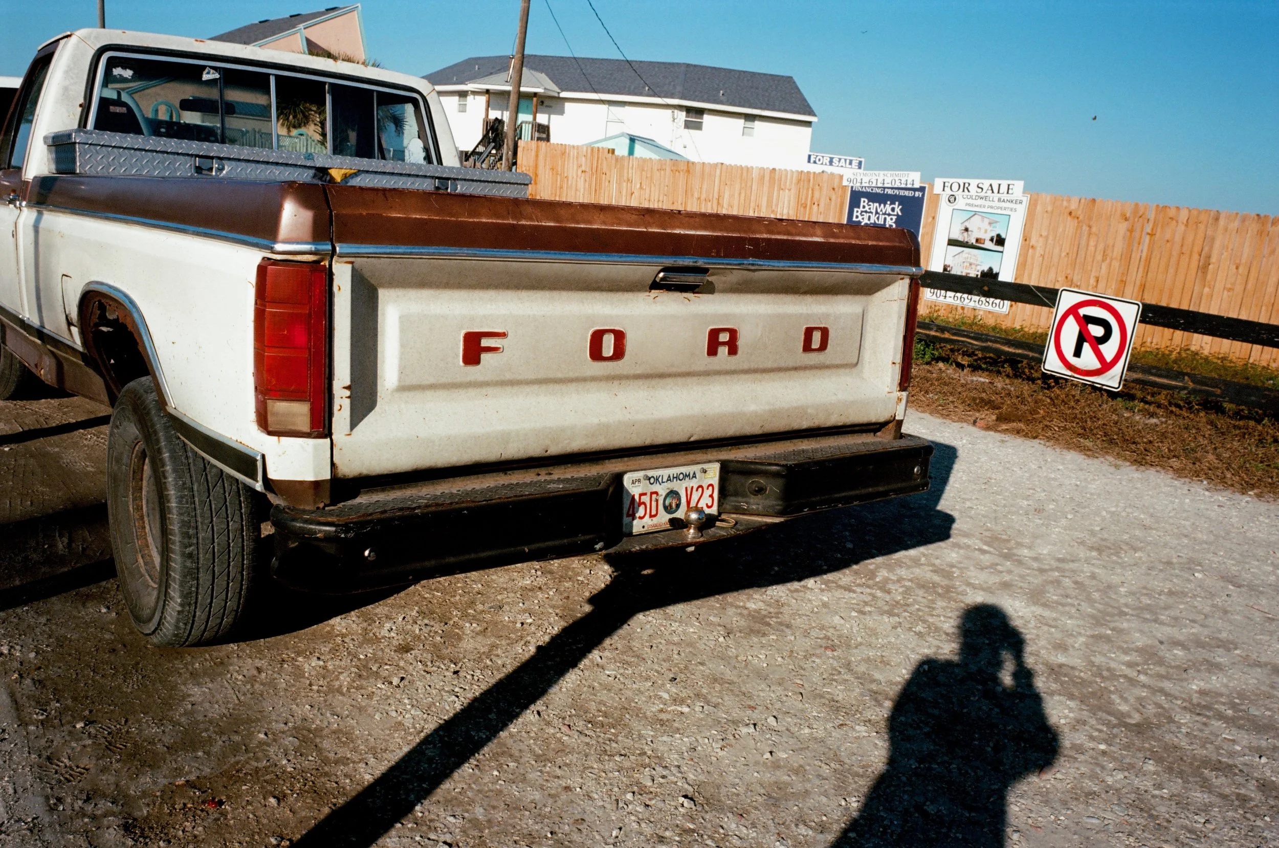 Old white Ford pickup truck with red letters on the tailgate parked on a gravel lot next to a wooden fence. There are "For Sale" signs in the background and a "No Parking" sign on the fence. The shadow of the photographer is visible in the foreground.