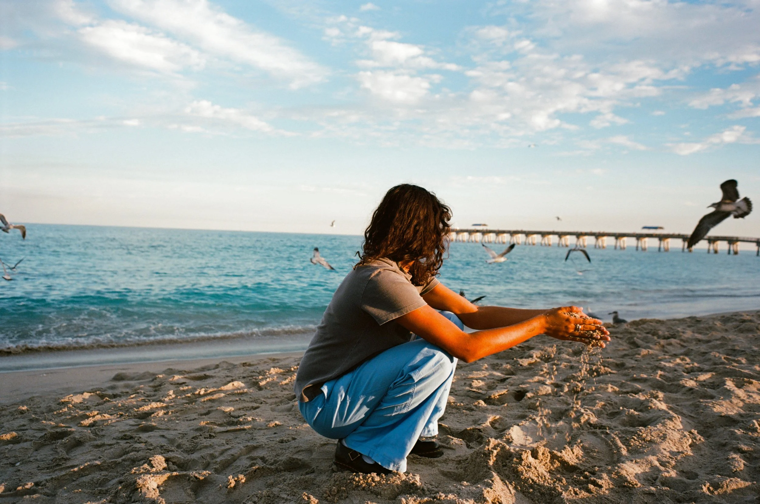 A woman with curly hair crouches on the sandy beach, pouring water from her hands, with seagulls flying in the sky and a pier extending into the ocean in the background.