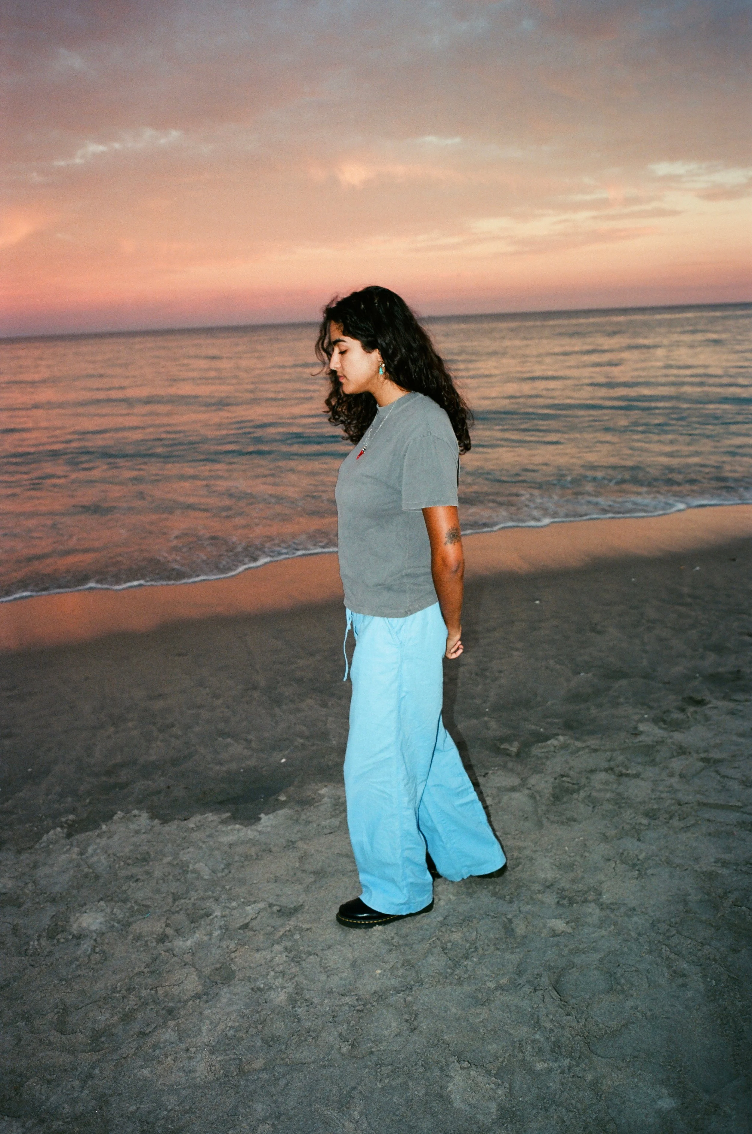 A woman with curly dark hair standing on a sandy beach at sunset, looking down, wearing a gray t-shirt and light blue pants.