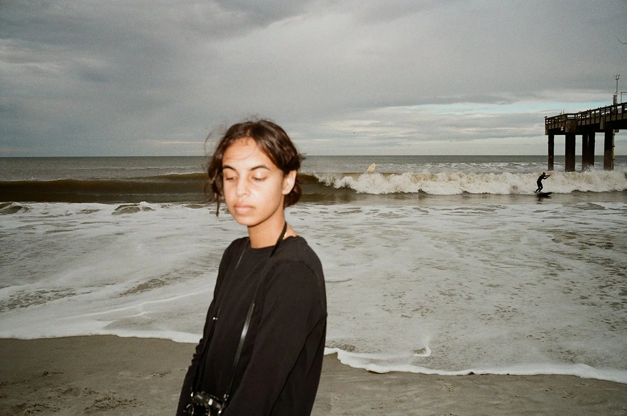 A young woman with short brown hair and closed eyes standing on the beach with ocean waves and a pier in the background.