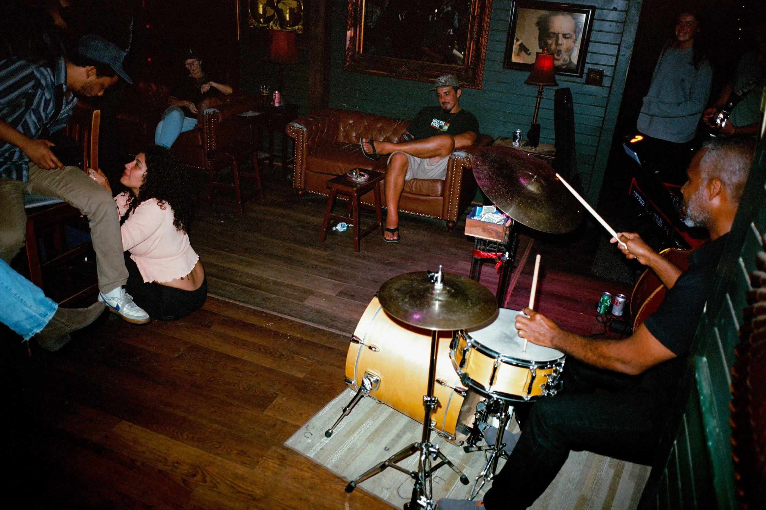 A man playing drums in a cozy, dimly lit lounge with people sitting on couches and chairs, a woman sitting on the floor talking to a man, and a woman standing in the background. The lounge has artwork on the walls and warm lighting.