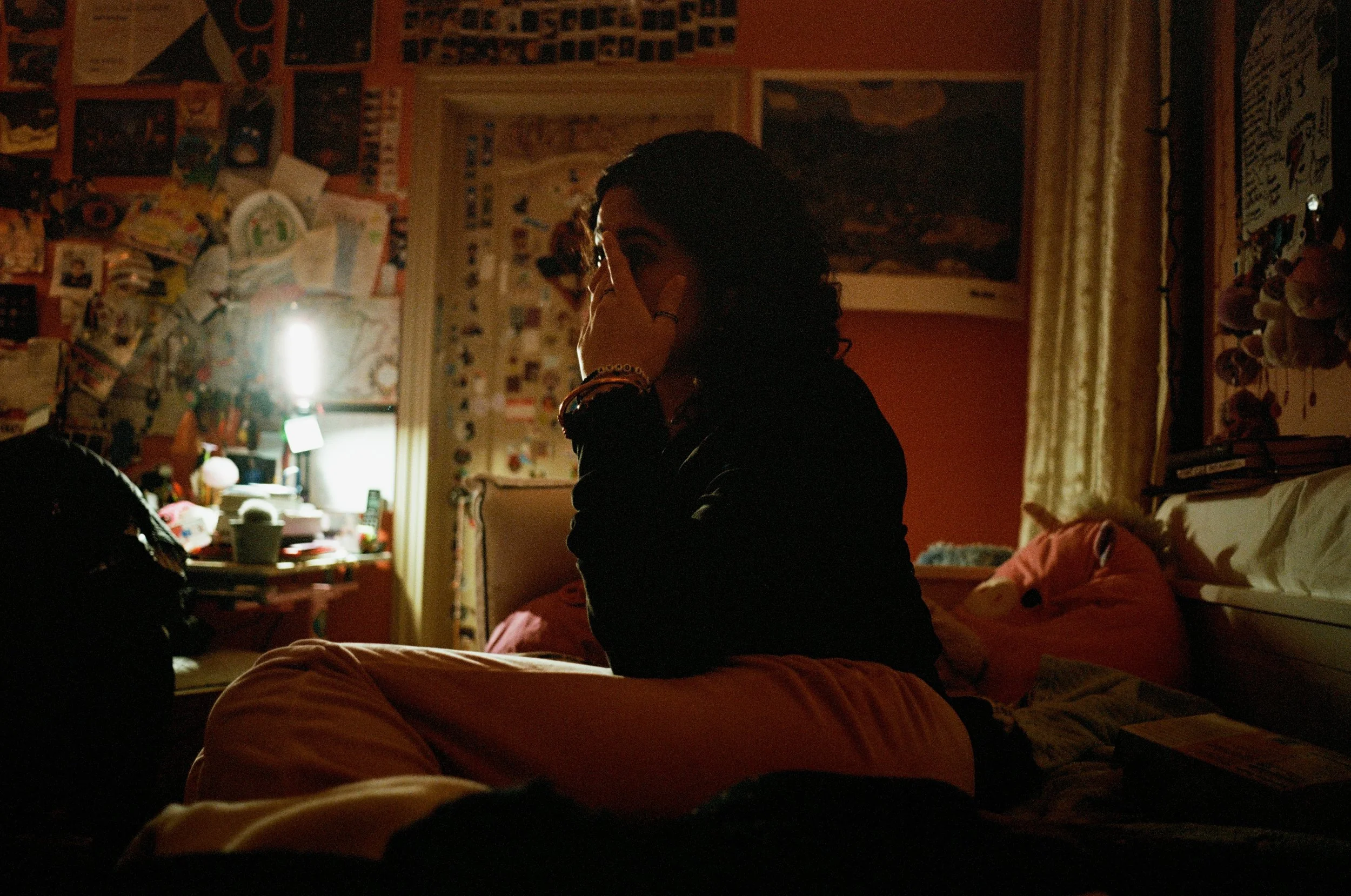 Person sitting on a bed in a dimly lit room, with various decorations, posters, and papers on the walls, and a window with a curtain in the background.