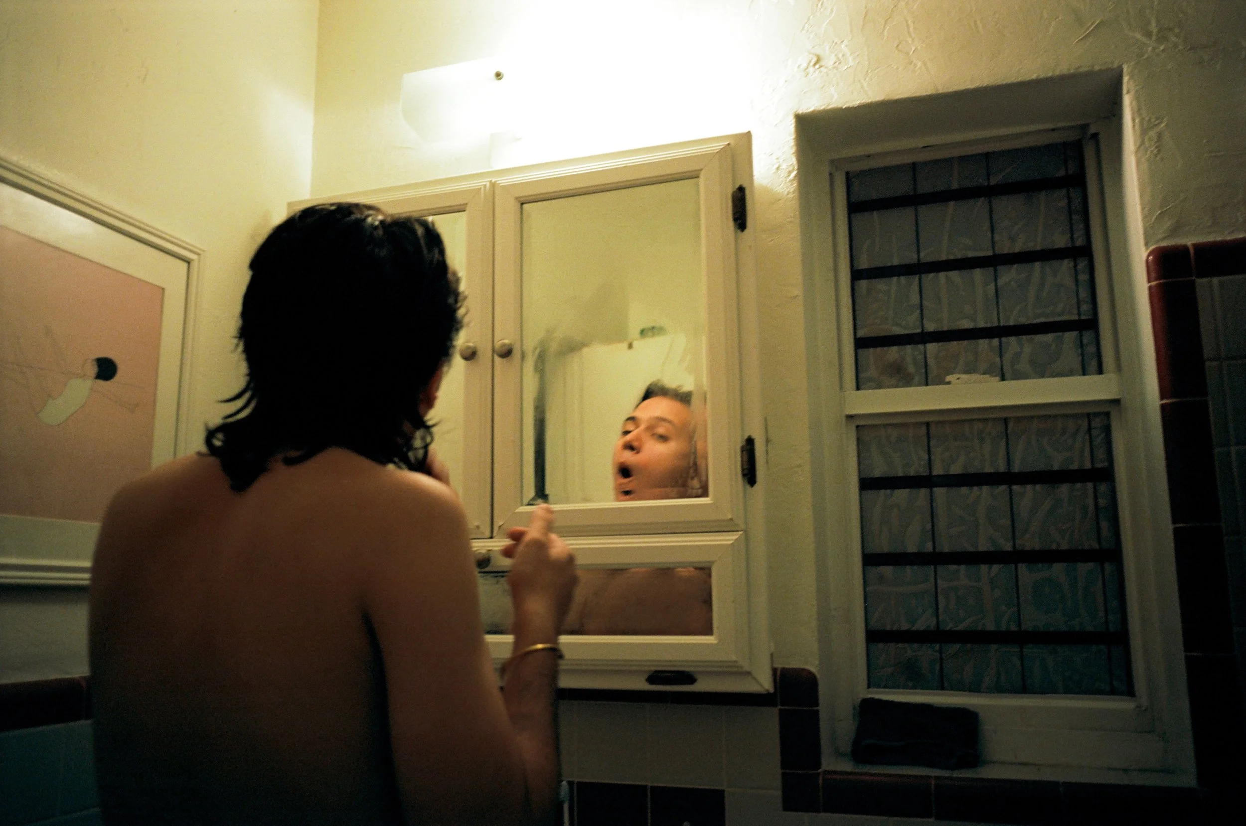 A person with short dark hair looking into a bathroom mirror, seeing their reflection with an exaggerated facial expression.