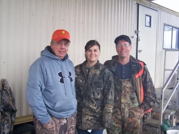 Three people standing outdoors in front of a beige building, dressed in casual and camouflage clothing, smiling at the camera.