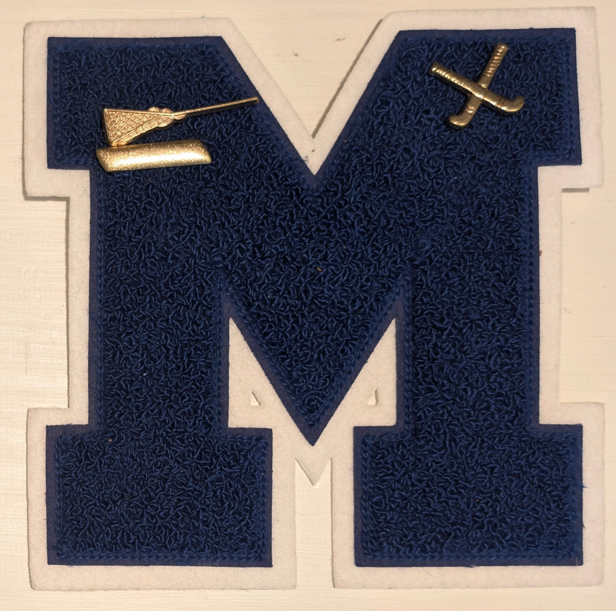 A large blue fabric letter M with white border and two small metallic golf club pins attached at the top left and right.