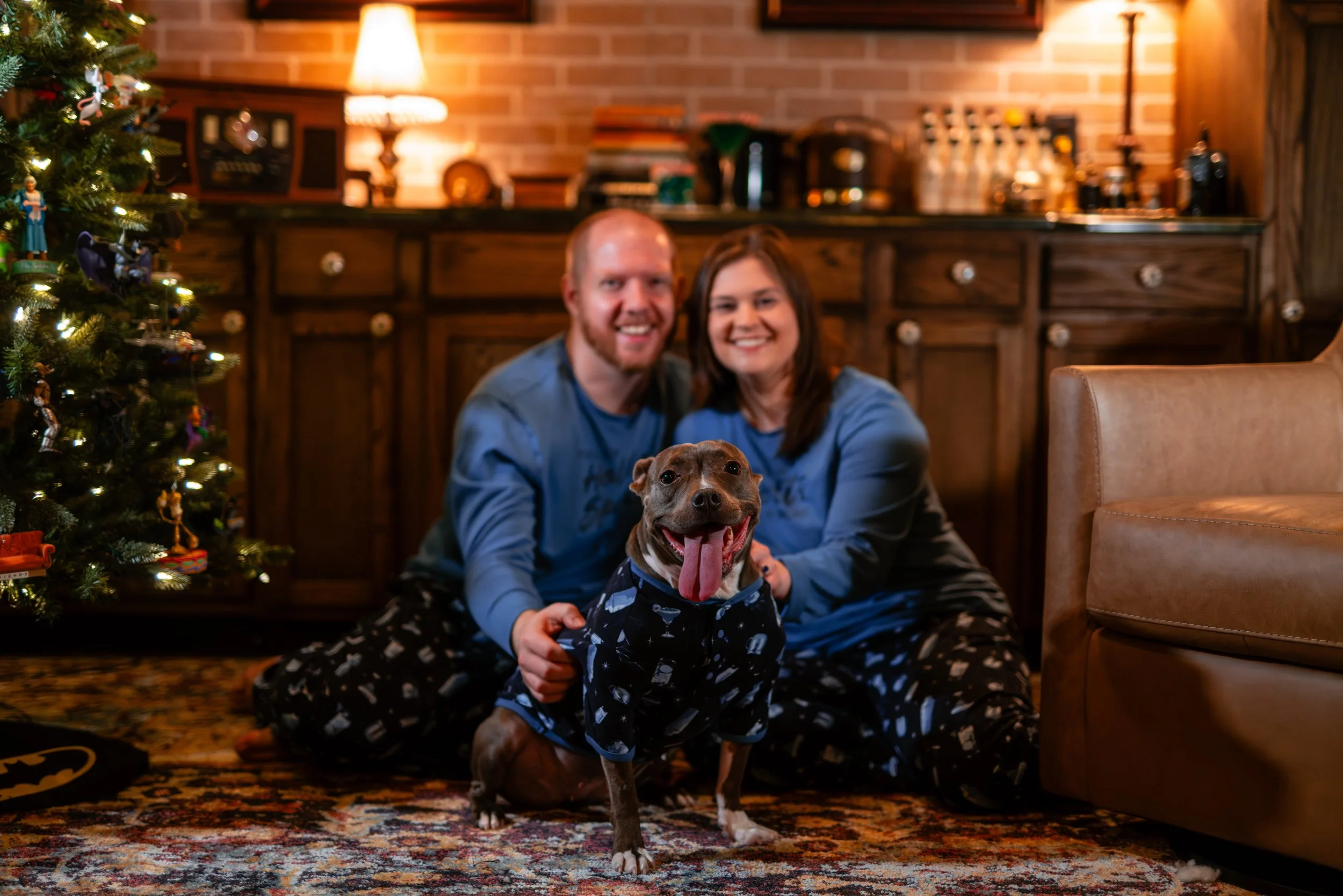 A happy couple sitting on the floor with their dog in front of a Christmas tree inside a cozy living room.