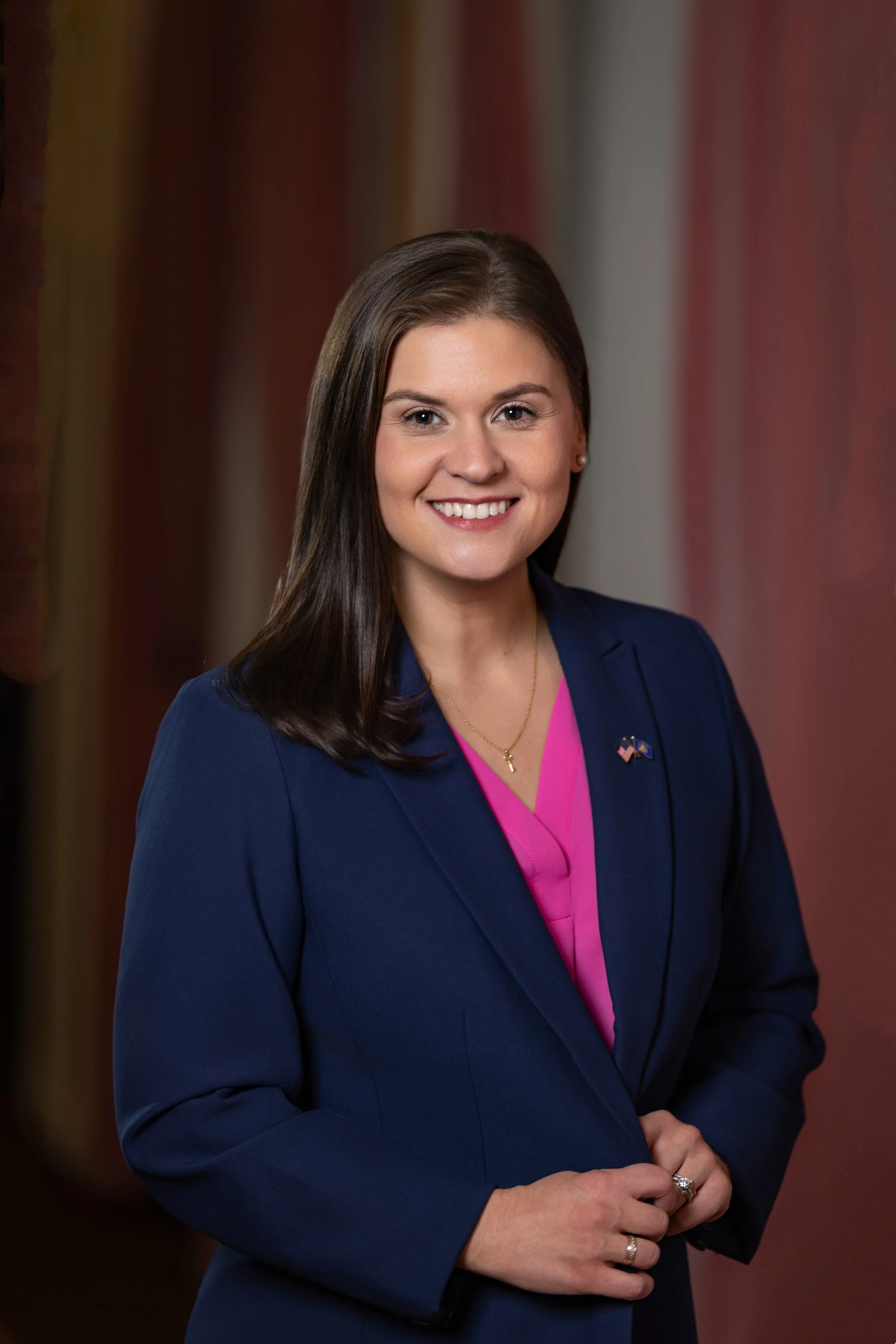 Kim Kohlhepp candidate for Pennsylvania State Representative. A professional woman with dark brown hair smiling, wearing a navy blazer and pink blouse with American flag pins, standing in front of a blurred background.