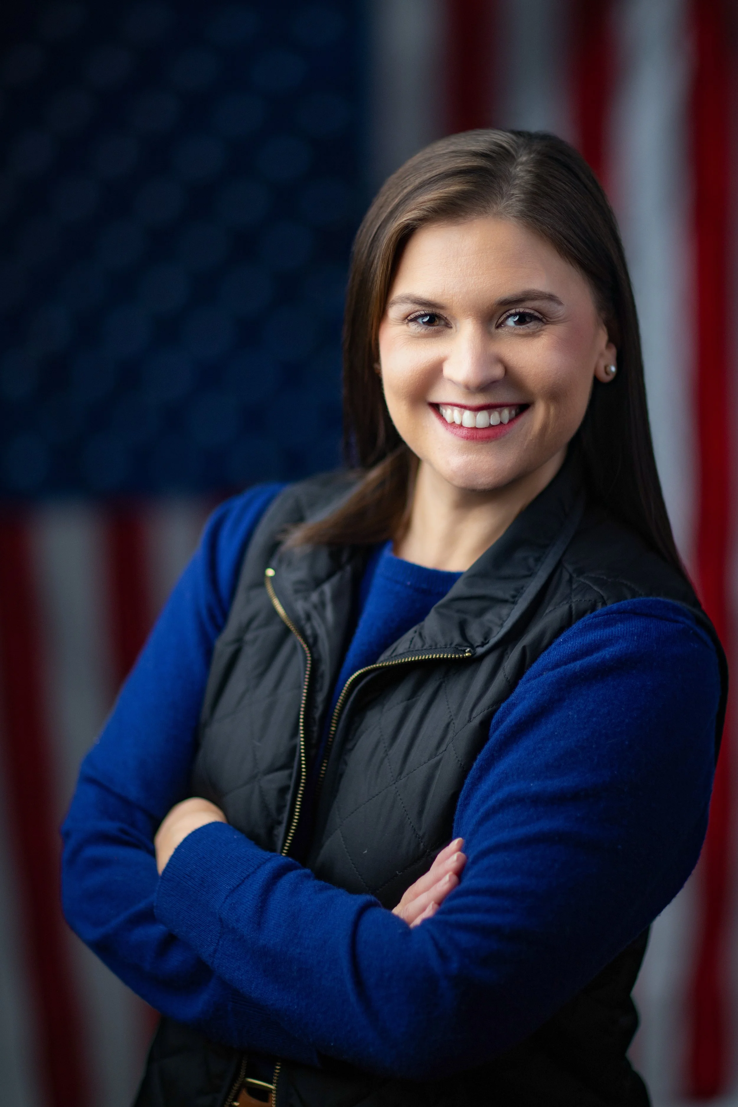 A woman with brown hair, smiling, wearing a blue sweater and a black vest, with an American flag background.
