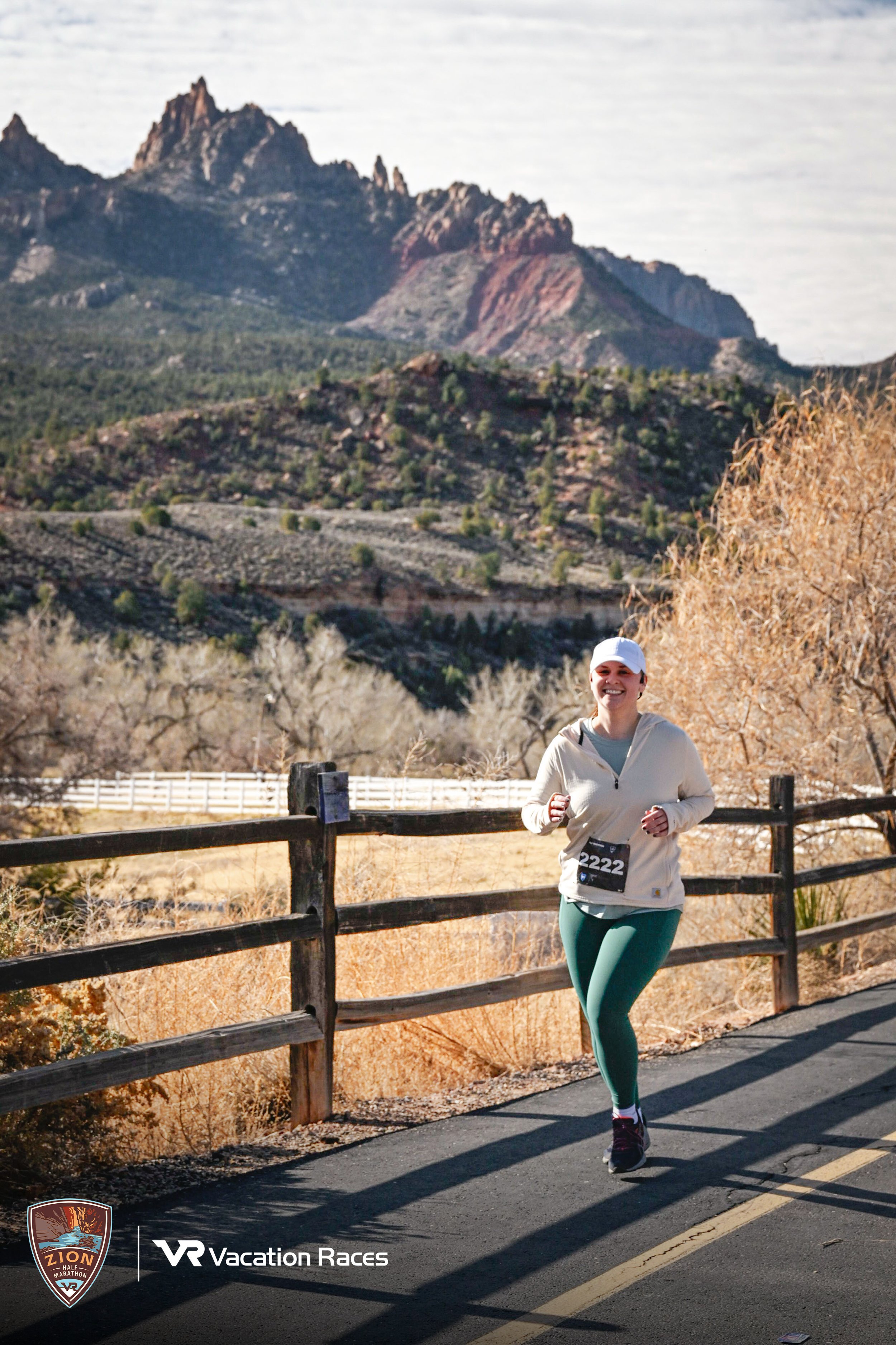 Kim Kohlhepp running a half marathon at Zion National Park. A woman wearing green leggings, a white jacket, and a white cap runs on a paved road during a race with a mountainous landscape in the background.