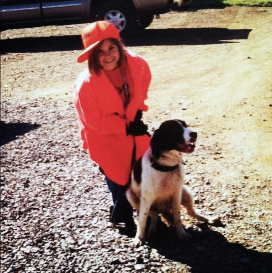 A young girl wearing an orange hat and a pink jacket, smiling and standing outdoors on gravel, with a black and white dog sitting beside her.