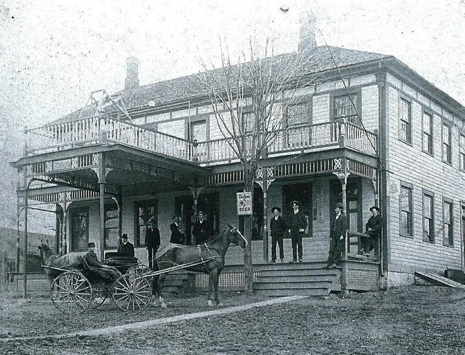 Black and white photo of a two-story wooden building with a wraparound porch. Several men in period clothing stand on the porch and steps. A horse and wagon are parked in front.