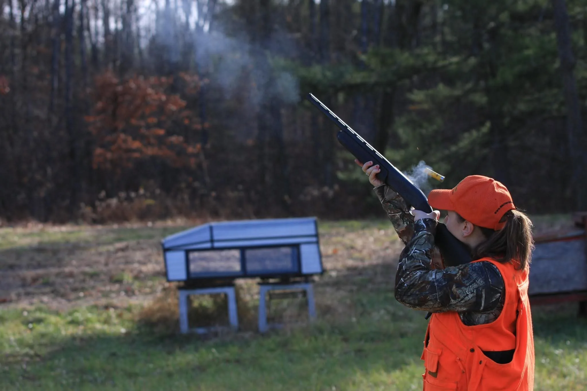 Young girl hunting with a shotgun in an outdoor setting, wearing an orange hunting vest and cap, with a forested background.