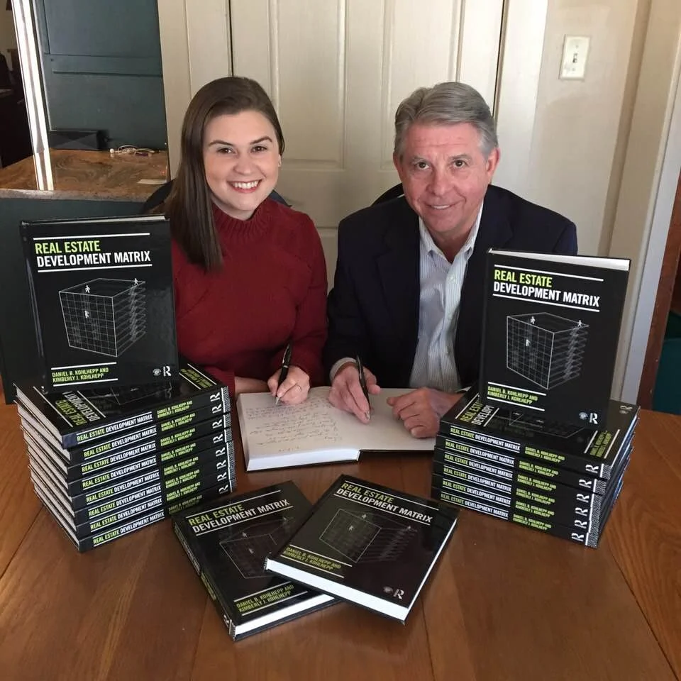 A man and woman sitting at a table with stacks of books titled 'Real Estate Development Matrix', signing papers. The woman is wearing a red sweater, and the man is wearing a dark suit jacket. They are smiling at the camera.
