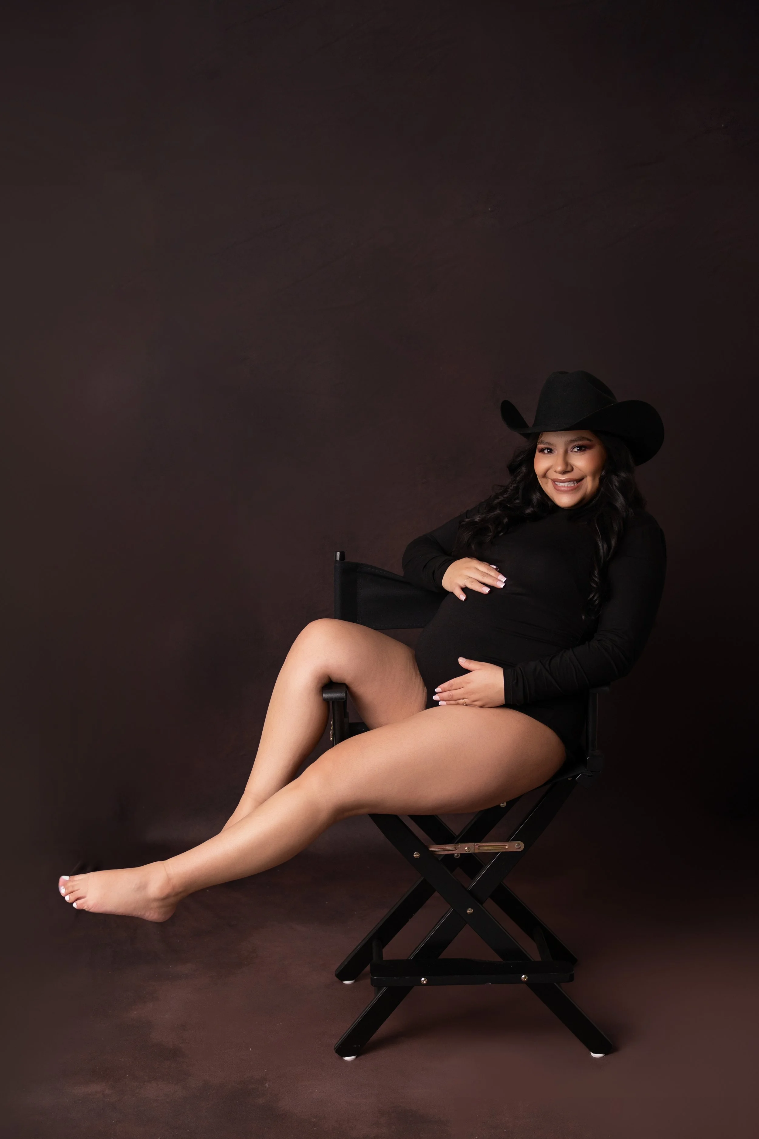 A woman in black clothing and a cowboy hat, sitting on a director's chair against a dark background, smiling and holding her pregnant belly.