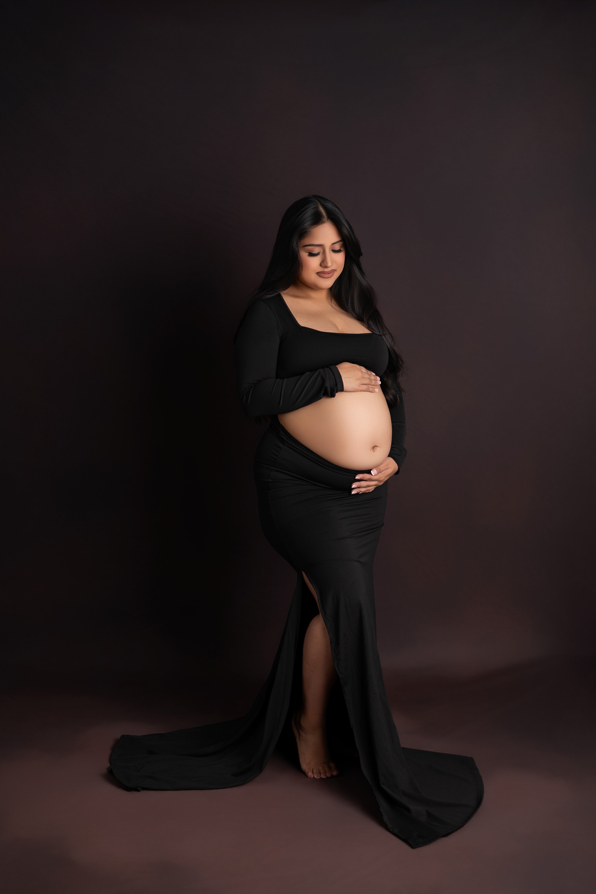 Pregnant woman in black dress standing against dark background with hands on her belly, smiling softly.