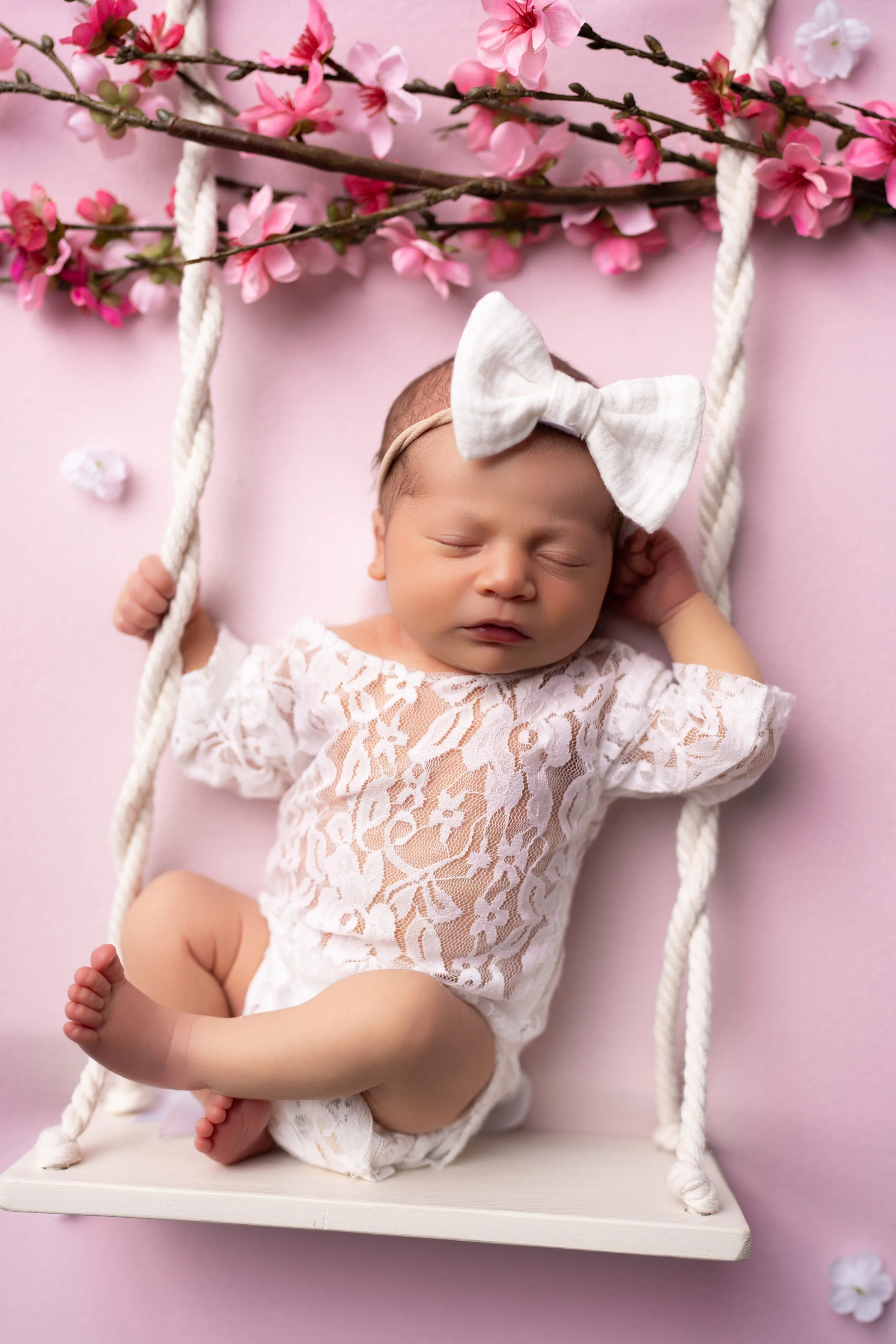 A baby girl sleeping on a white swing with pink flowers and branches overhead, dressed in a white lace outfit and a white bow headband.