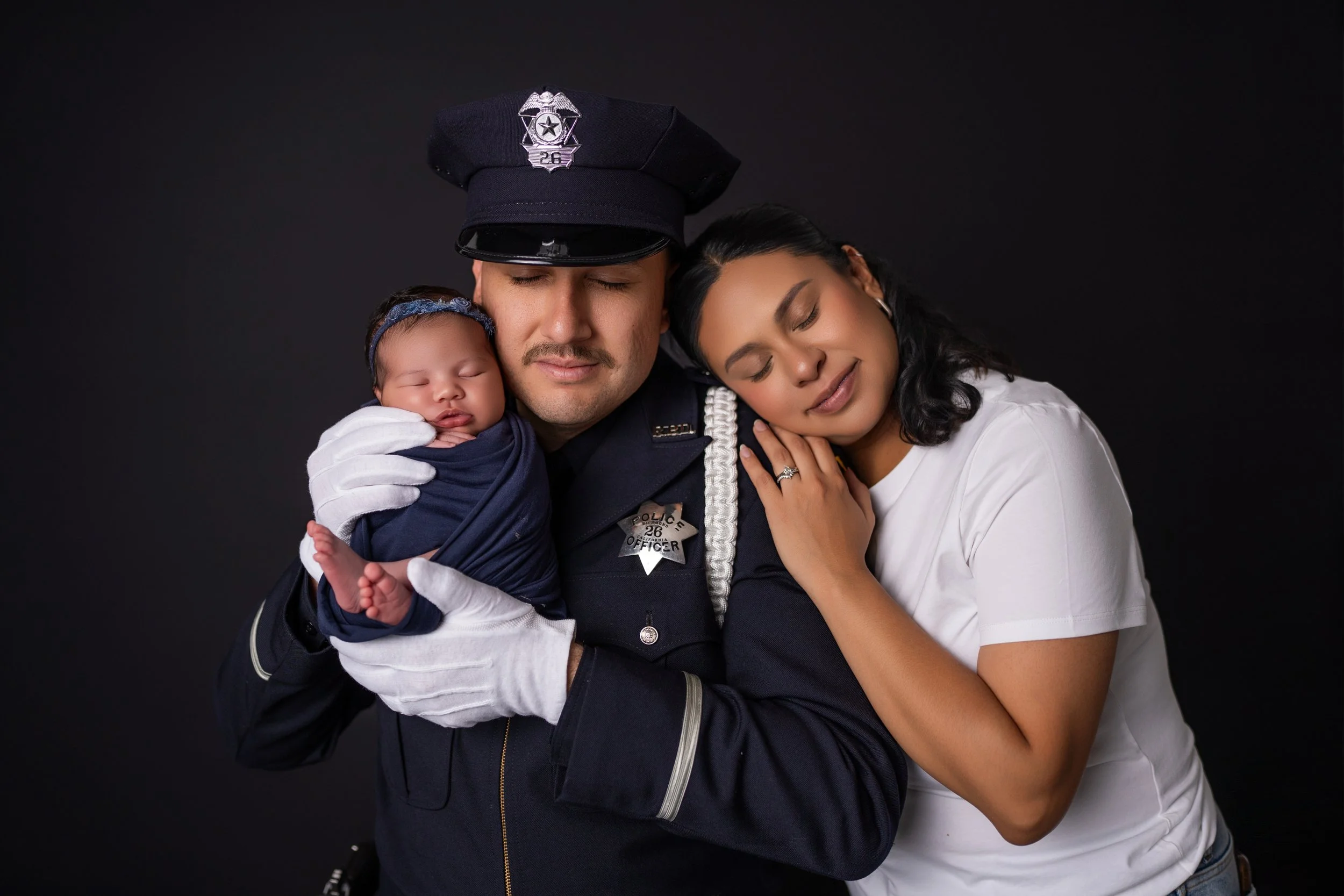 A police officer in uniform holding a sleeping baby, with a woman resting her head on his shoulder, all smiling against a black background.
