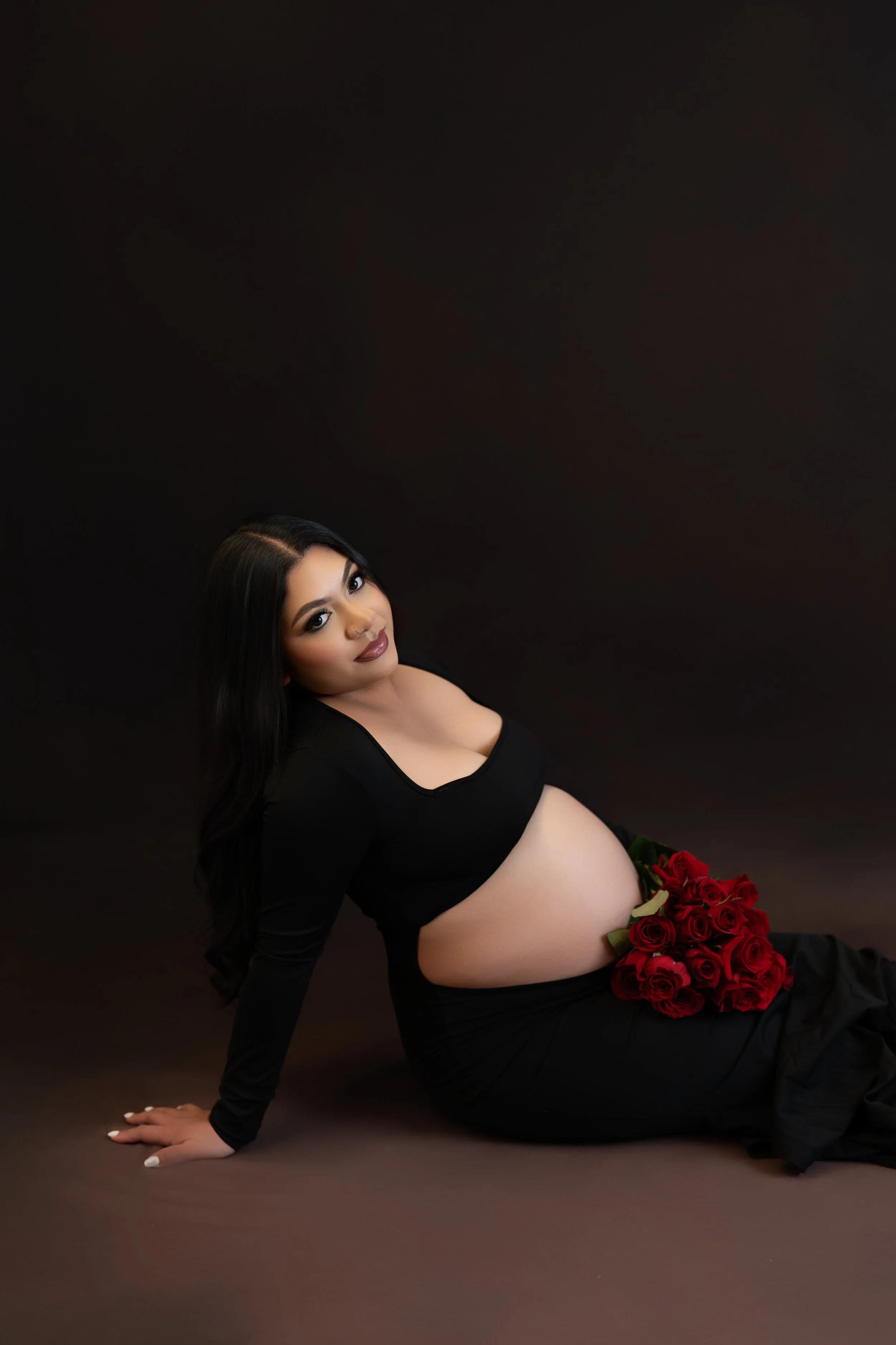 A pregnant woman with long dark hair and makeup, wearing a black top and black skirt, sitting on the floor against a dark background, holding a large bouquet of red roses resting on her baby bump.