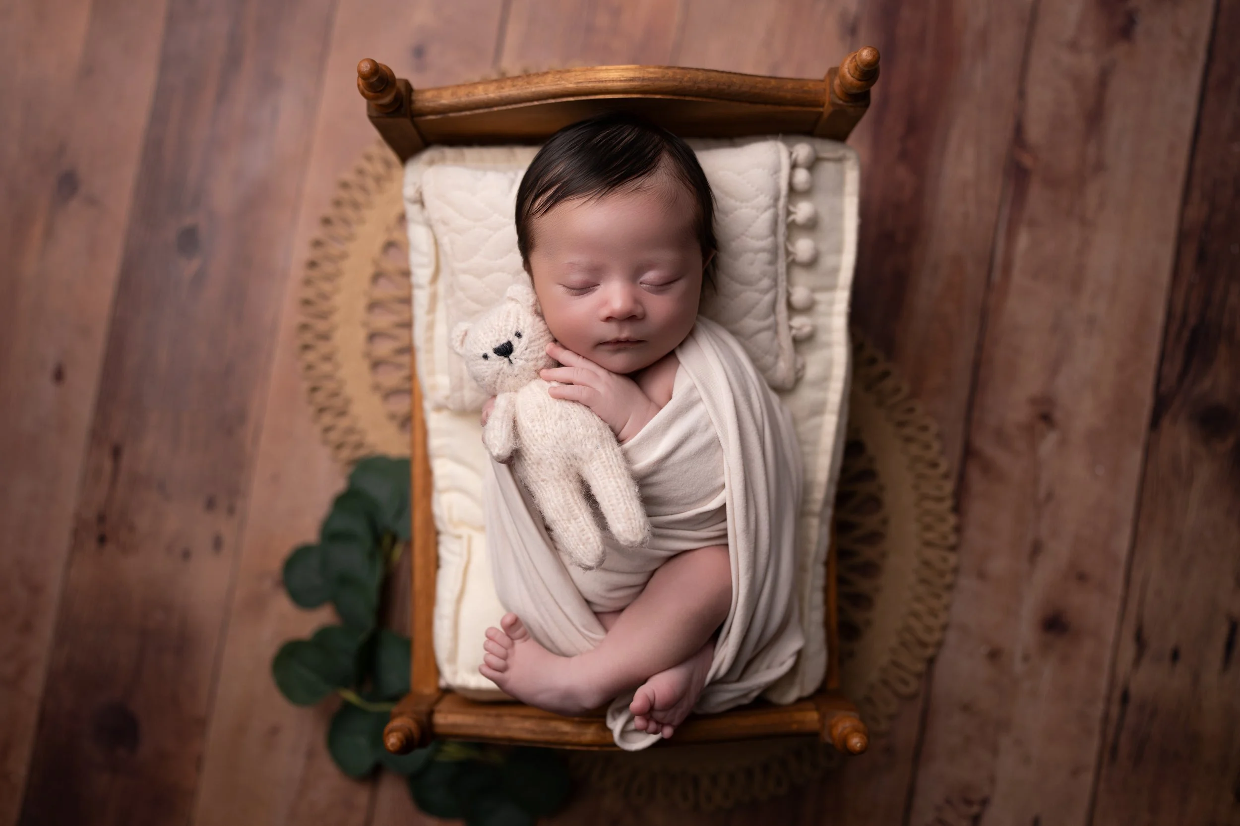 A newborn baby peacefully sleeping on a small wooden bed, hugging a small knitted teddy bear, with a beige blanket and a pillow. The bed is on a wooden floor, and there is a small green plant nearby.