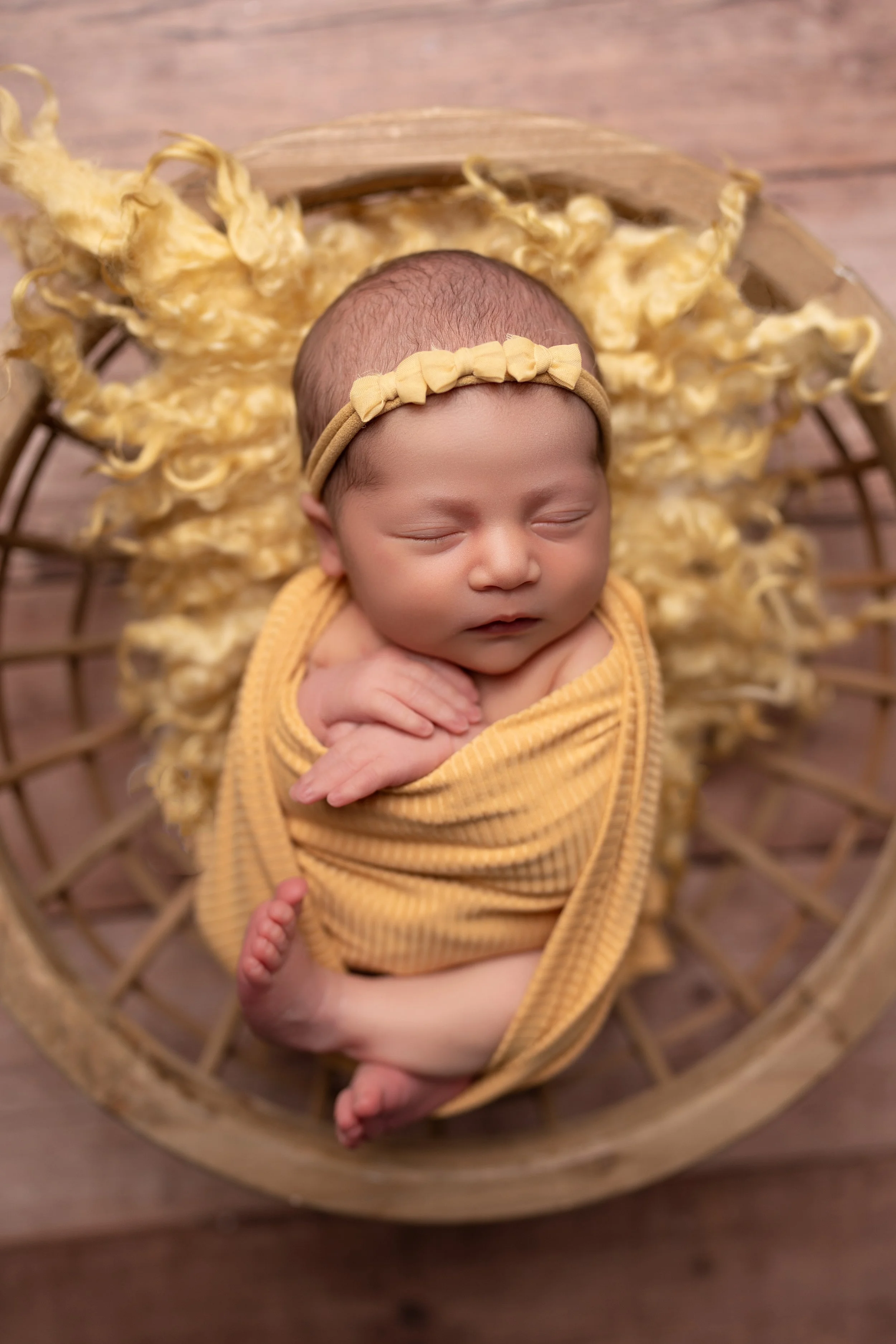 Swaddled newborn baby resting in a round wicker basket on a wooden floor, with curly yellow blanket underneath, wearing a matching yellow headband.