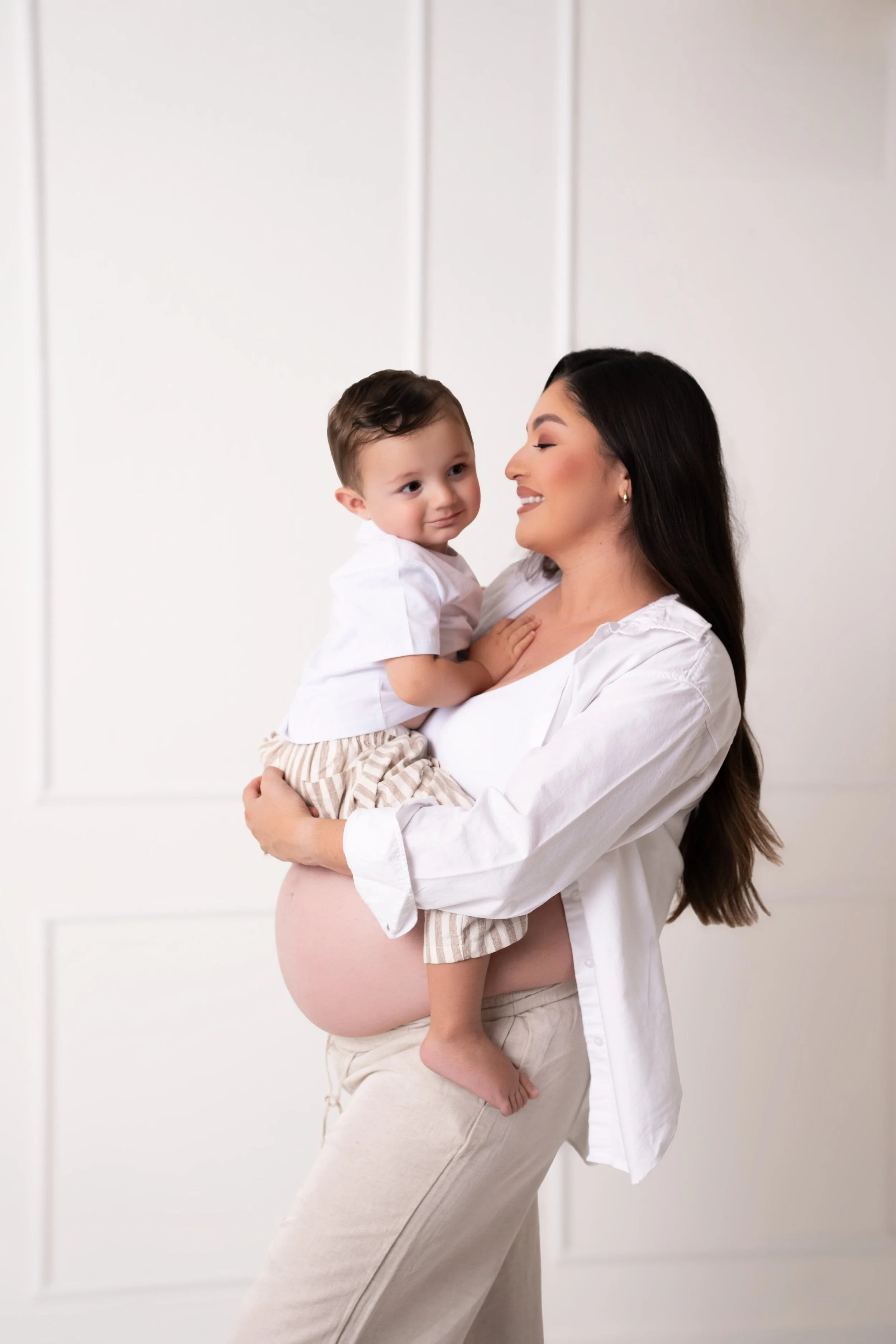 A pregnant woman holding a young boy, smiling and looking at him, in a bright white room.