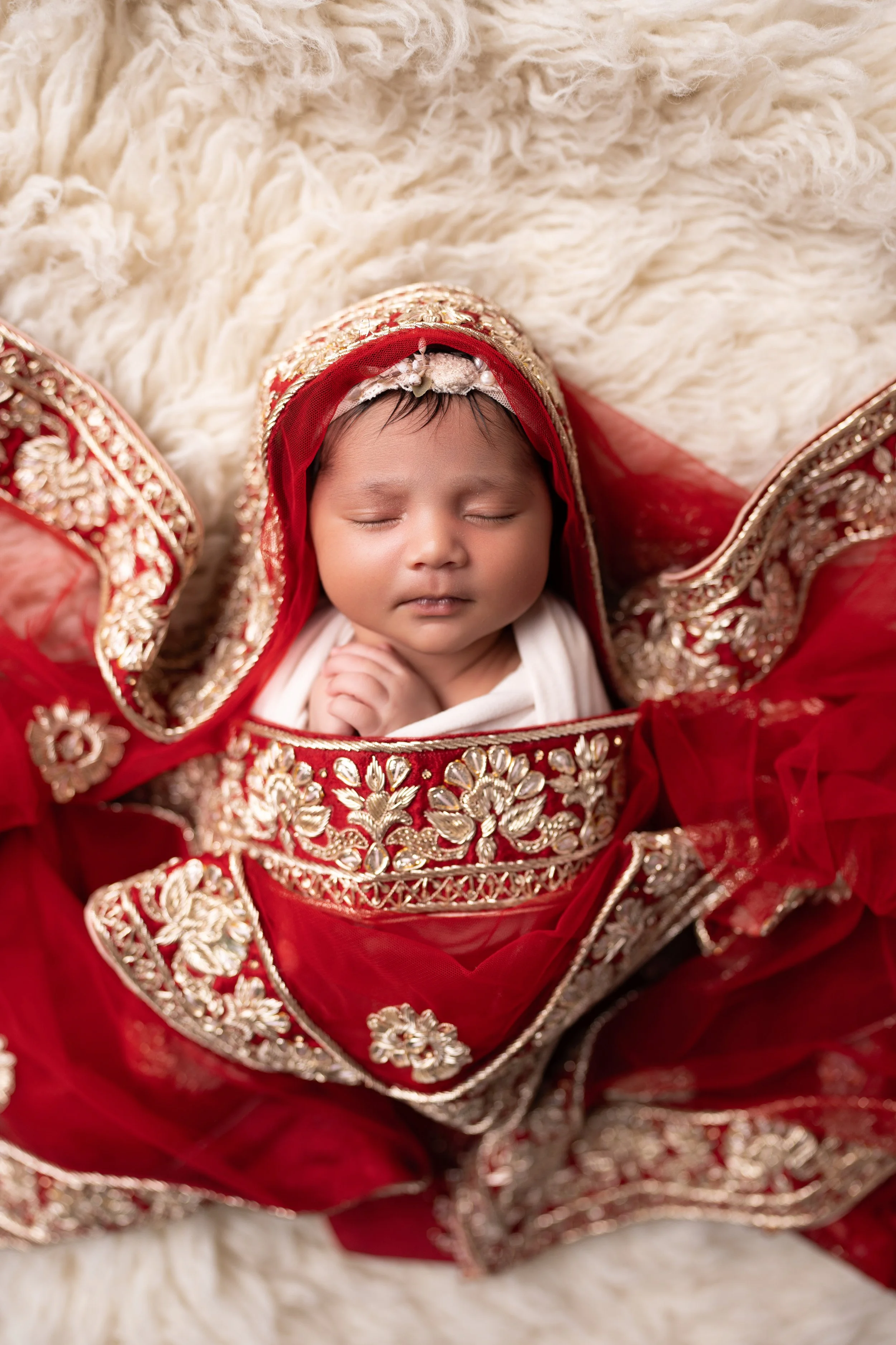 A sleeping baby dressed in traditional Indian attire with intricate gold embroidery and red fabric, lying on a fluffy cream-colored surface.