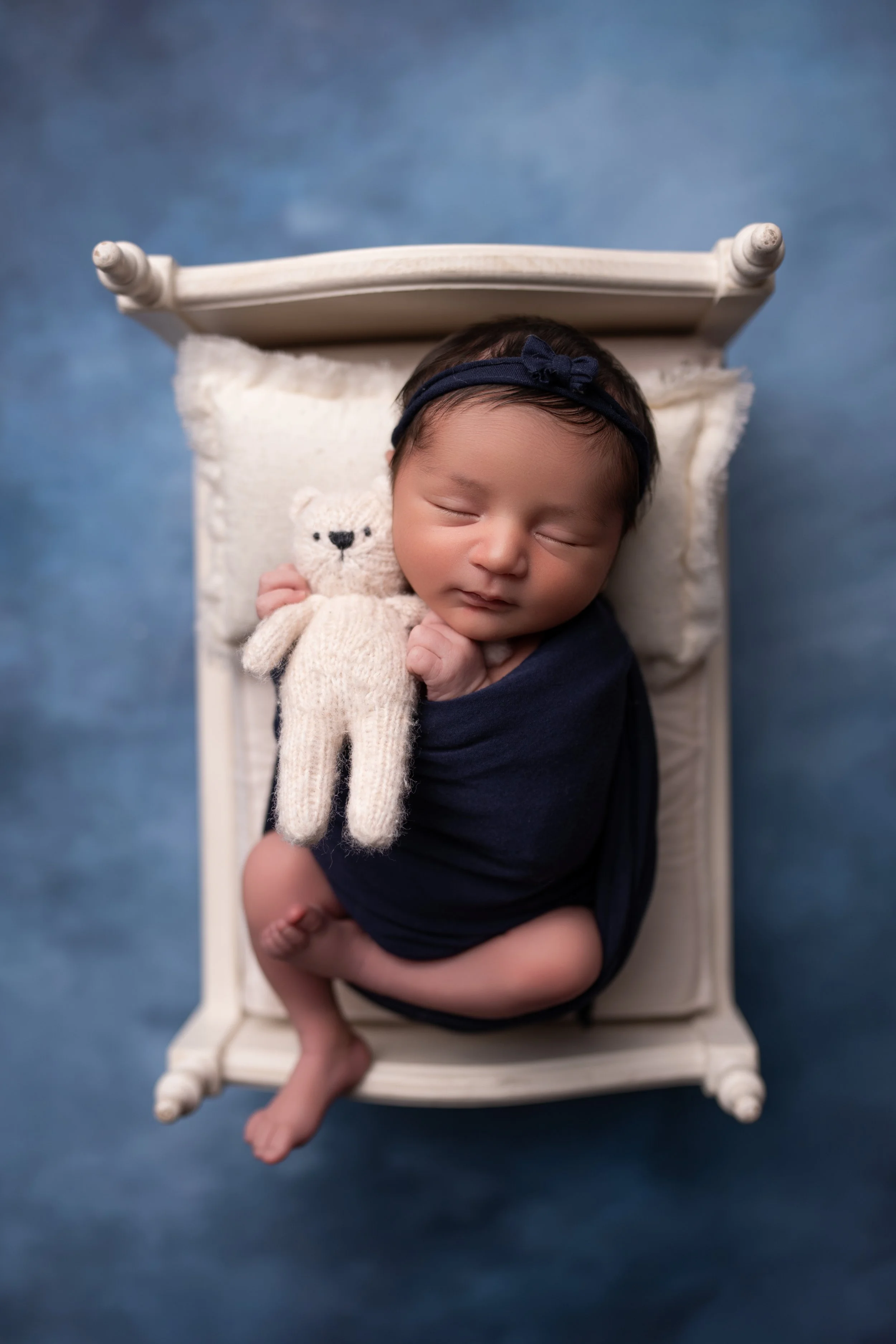 Newborn baby sleeping on a small bed, holding a cream-colored teddy bear, wrapped in a dark blanket, with a dark headband.