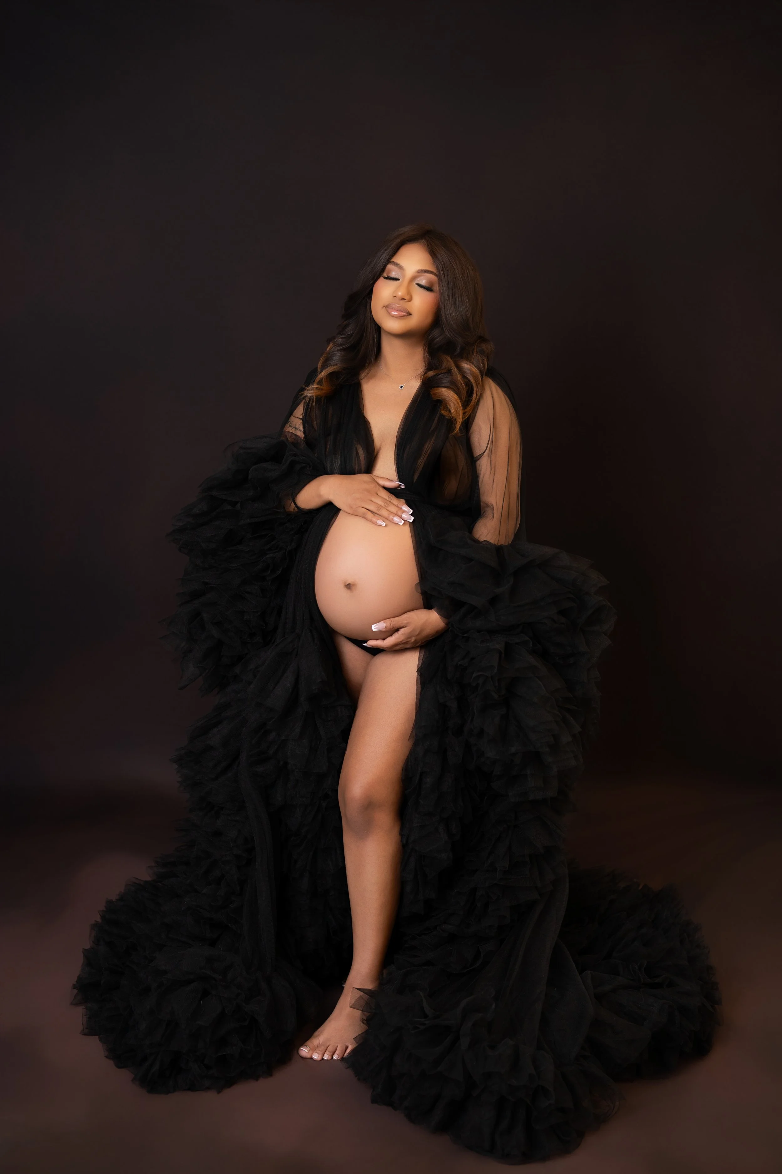 A pregnant woman with long wavy hair wearing a black sheer gown with ruffled details, posing against a dark background.