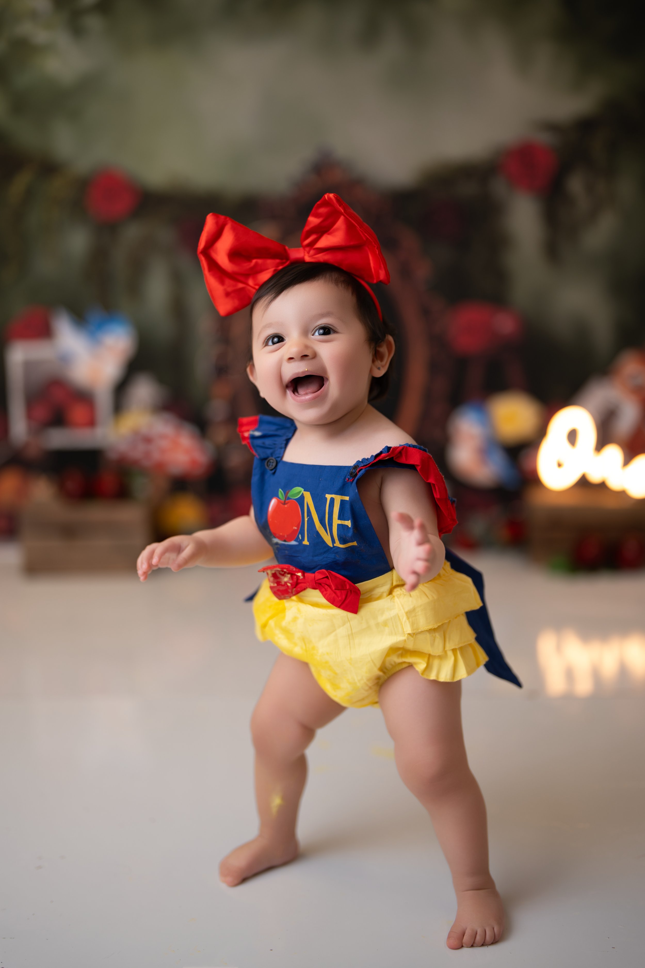 Adorable baby in Snow White costume with a red bow, blue top with a red apple and yellow skirt, smiling and standing with arms out, in a festive decorated setting.
