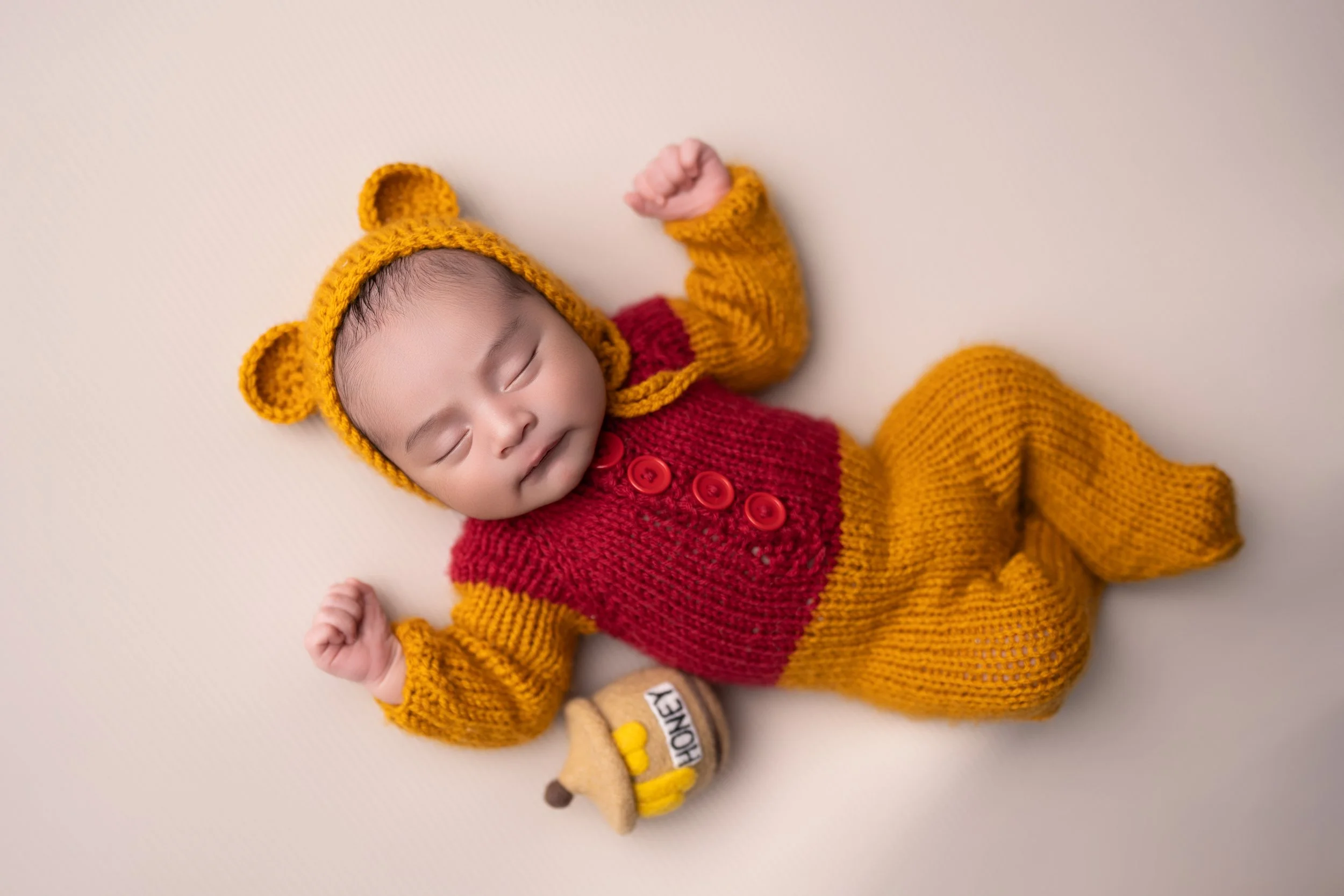 A sleeping baby wearing a yellow bear hat, a red and yellow knitted outfit, and holding a small plush honey jar, lying on a cream-colored surface.