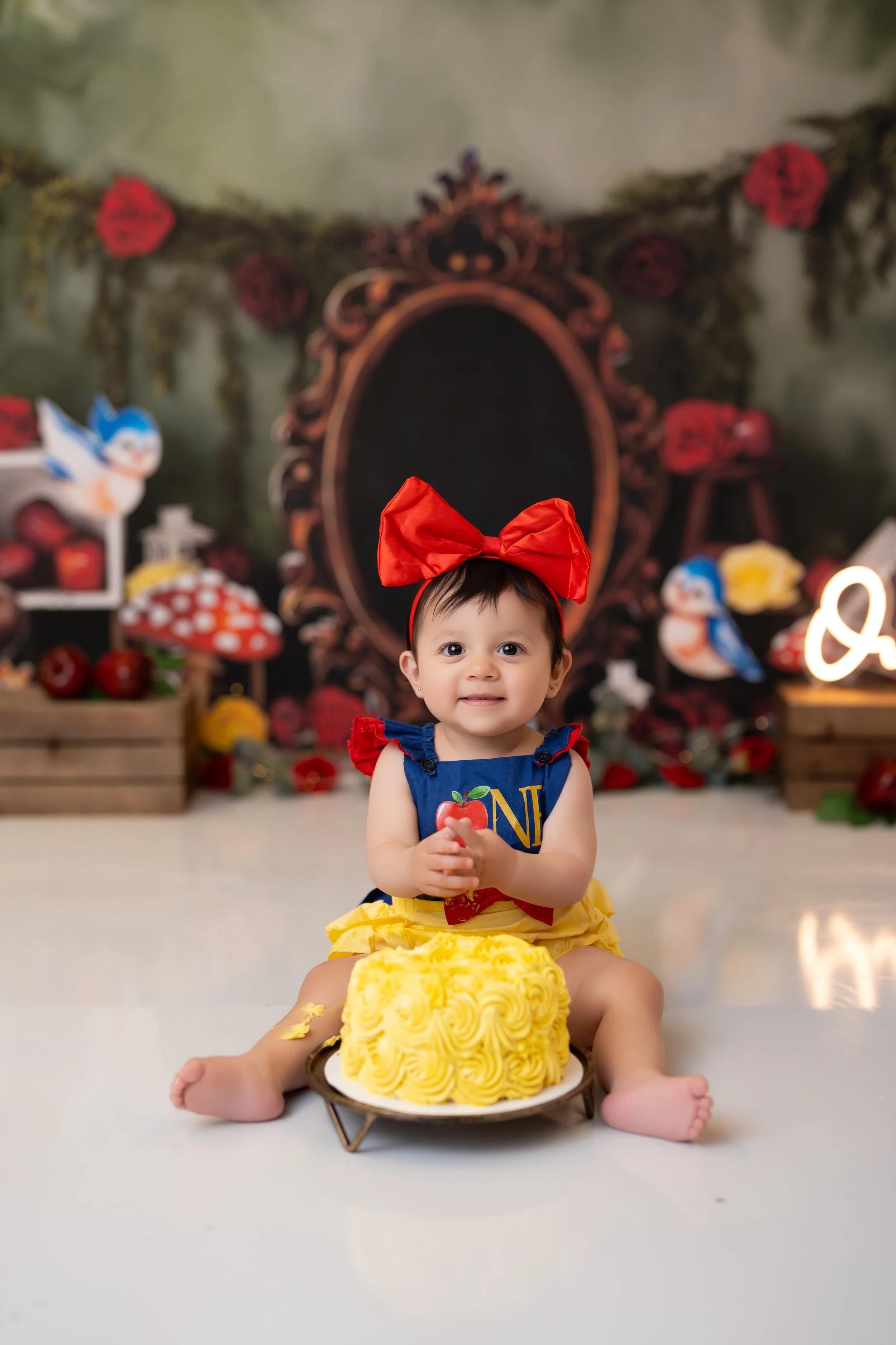 A young girl in a Snow White costume with a red bow on her head sitting on the floor in front of a yellow birthday cake with swirled icing. She is holding a small object and smiling, with a decorated backdrop featuring fairy tale and woodland element