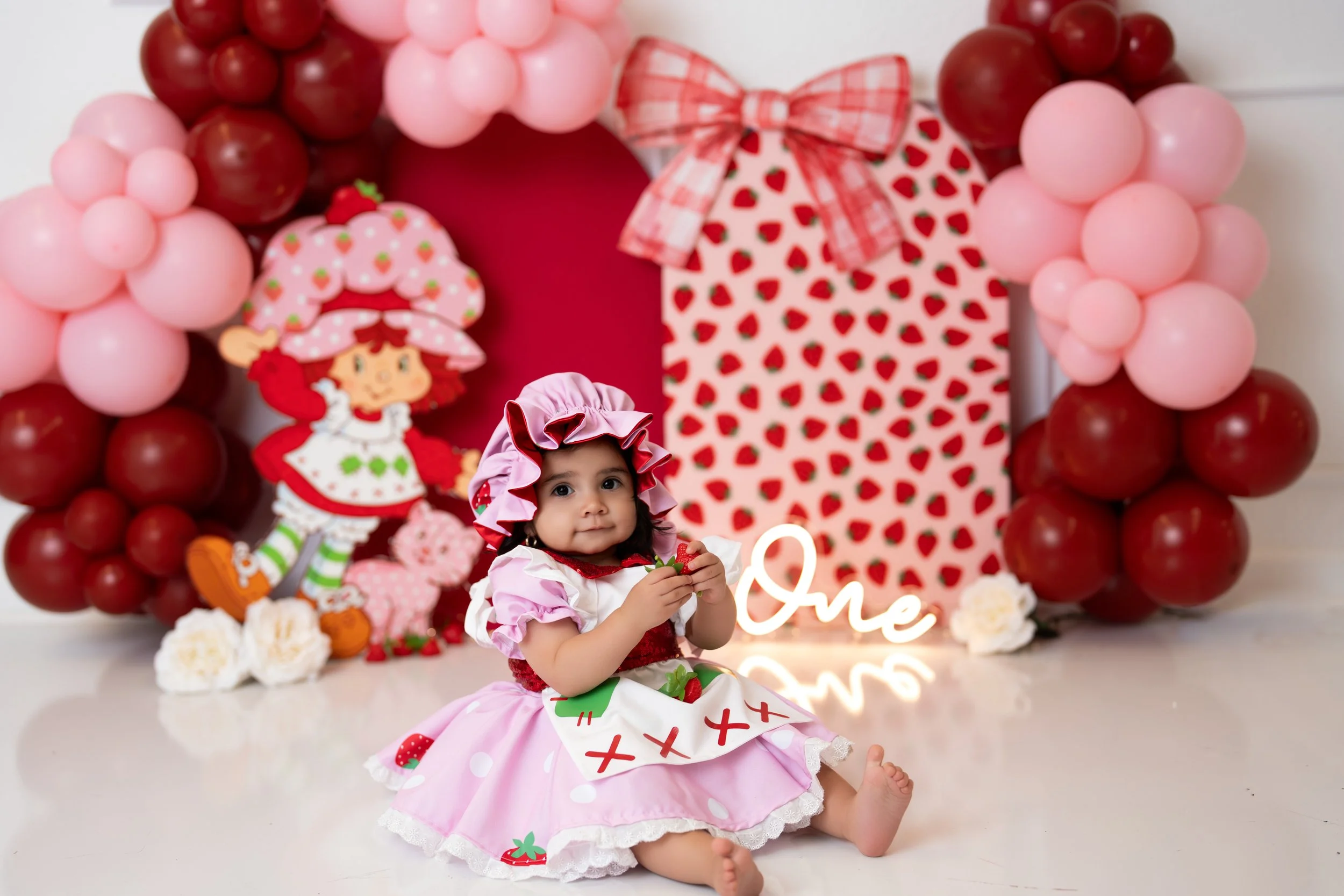 A young girl dressed in a strawberry-themed costume, sitting on the floor holding a strawberry, with a decorated child's birthday backdrop featuring pink and red balloons, a strawberry-themed cutout, and a neon sign that says "One".
