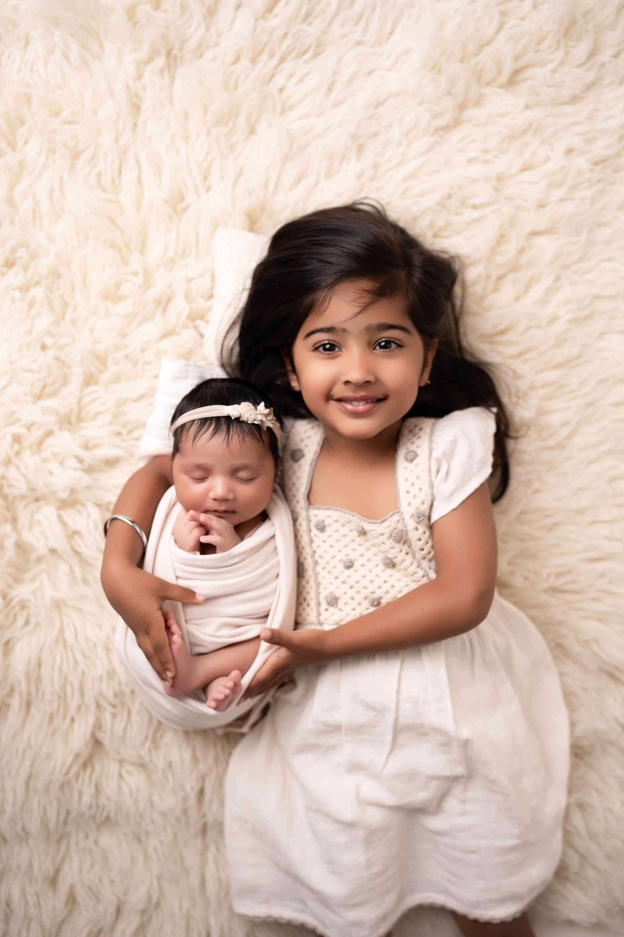 Two young girls, one older and one younger, lying on a fluffy cream-colored blanket. The older girl is smiling and has dark hair, while the younger girl is sleeping with a headband. The older girl is holding the younger girl gently.