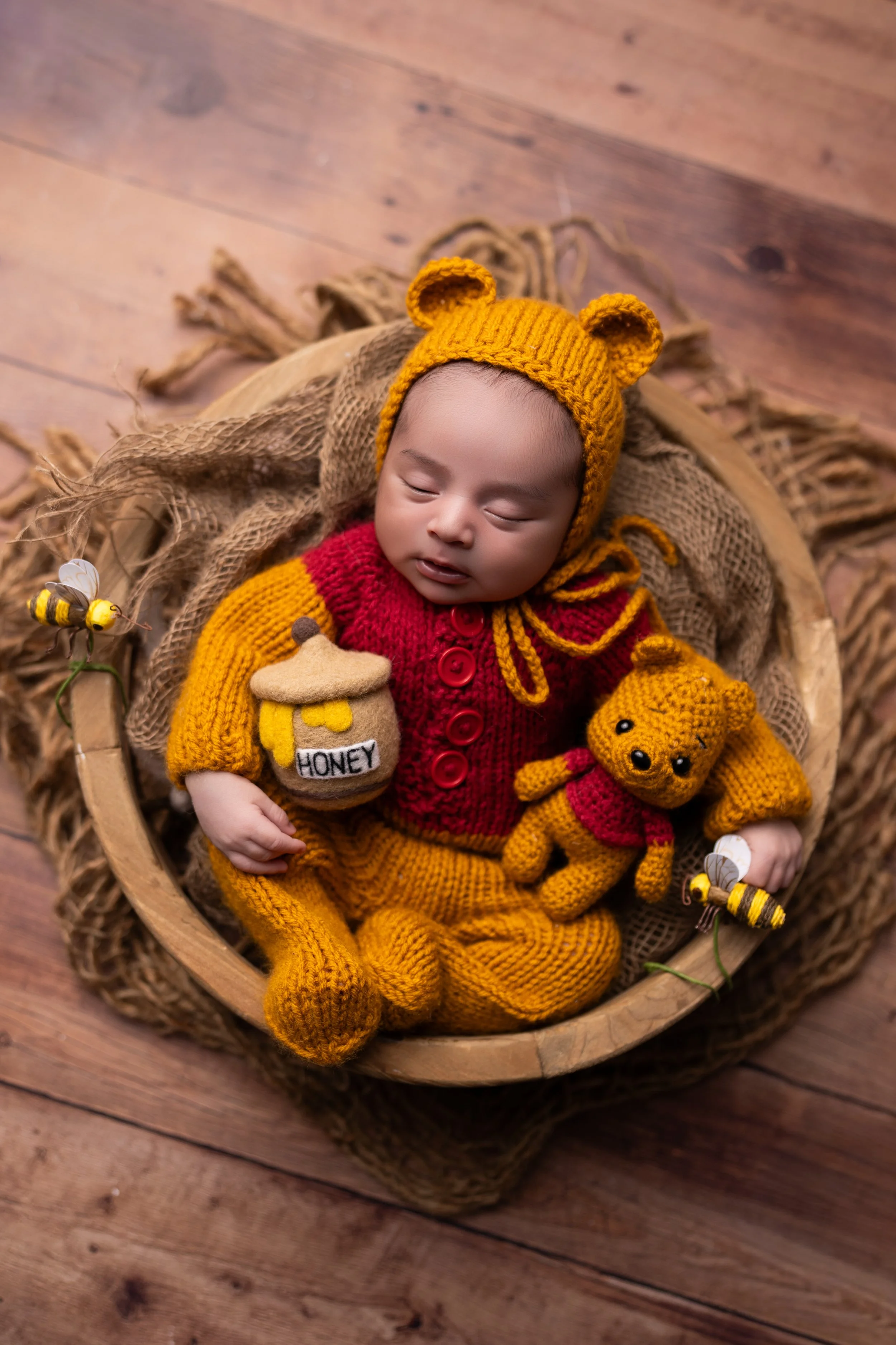 A sleeping baby dressed in a yellow bear hat, red and yellow knitted outfit, lying in a basket with stuffed bear and honey jar toys, surrounded by decorative bees and flowers.