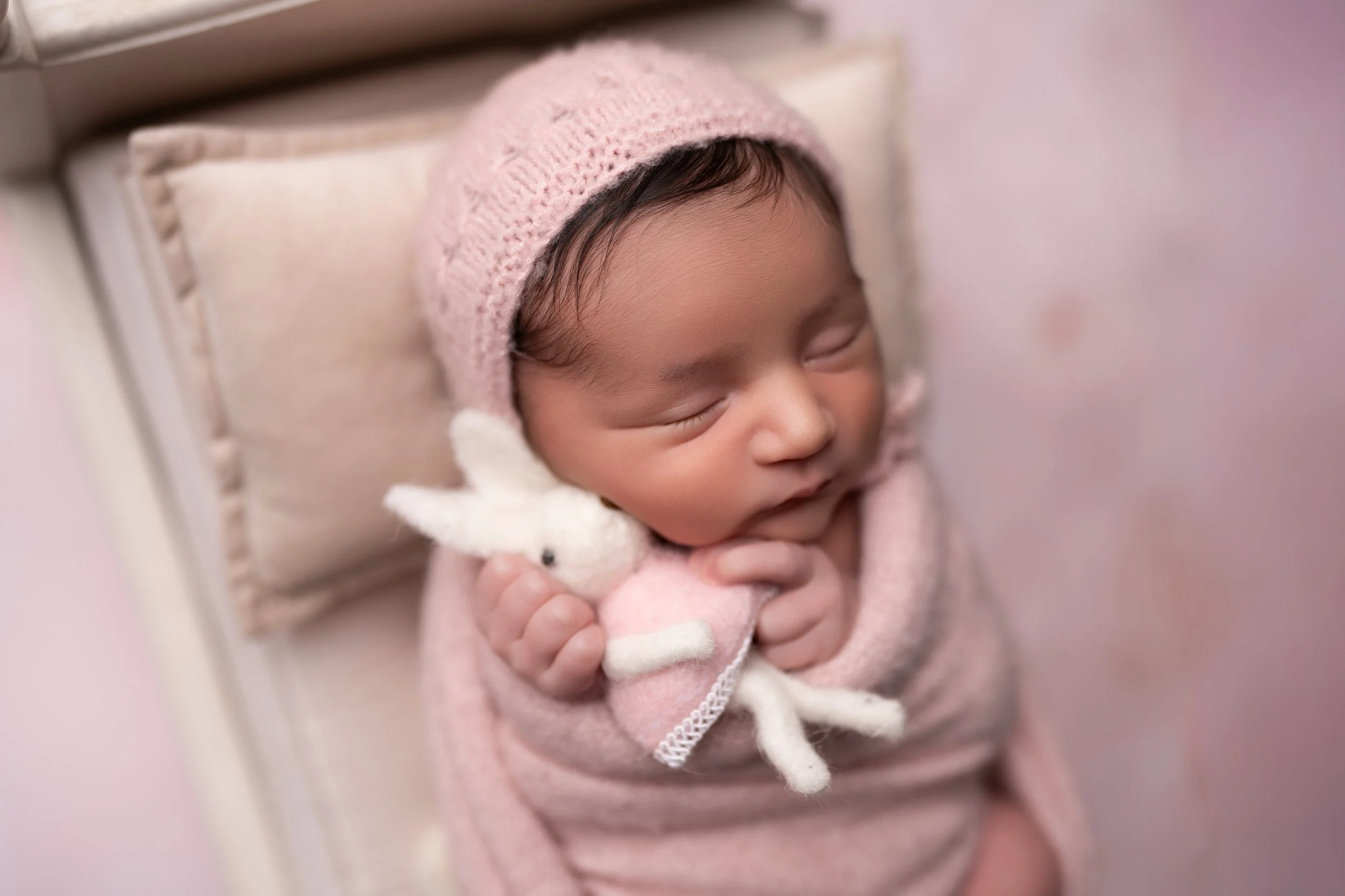 A sleeping baby wearing a pink knit hat, holding a small white stuffed bunny, and wrapped in a pink blanket.