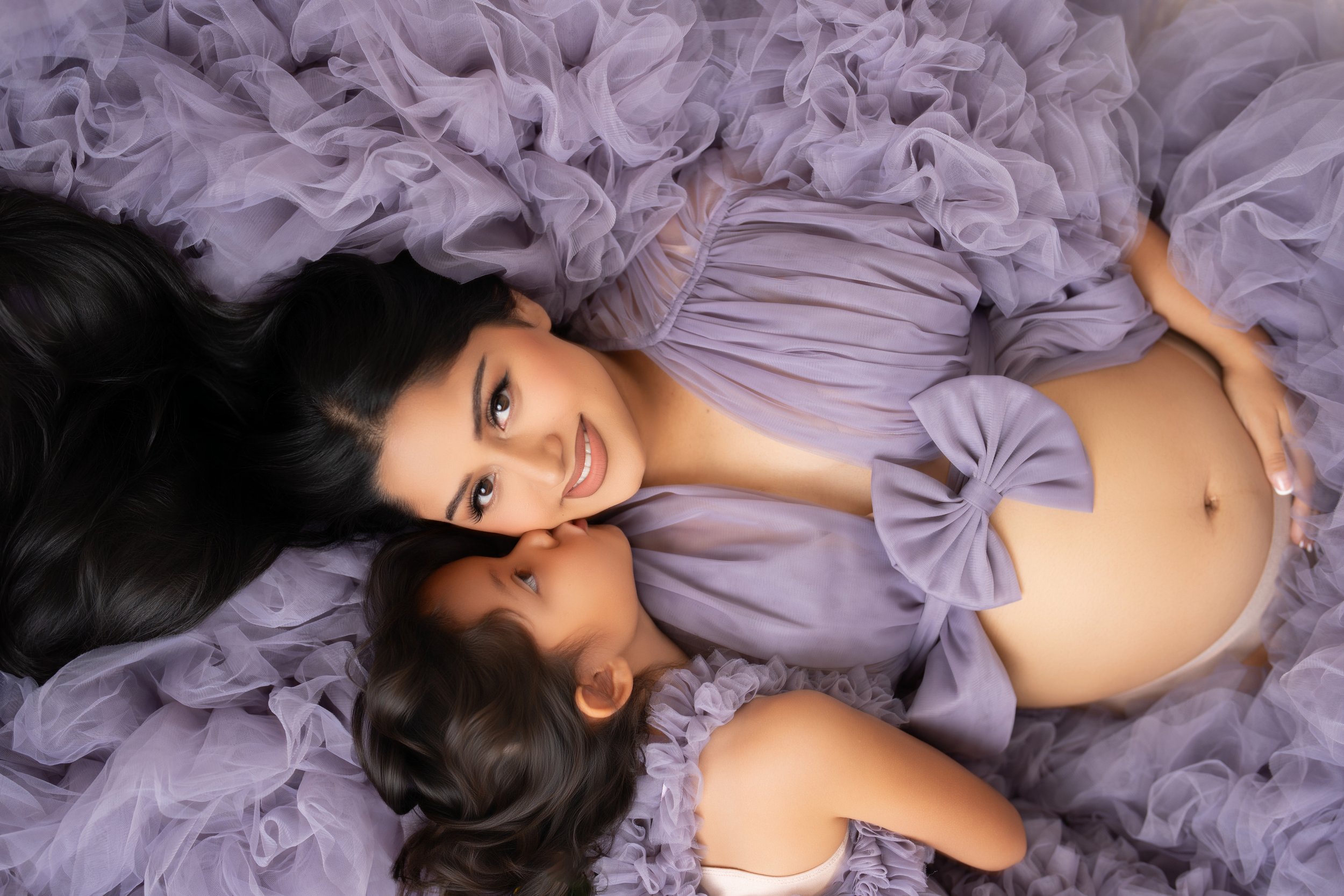 A young woman lying on purple ruffled fabric with a young girl, both wearing matching purple outfits with bows, and the girl resting her head on the woman's shoulder.